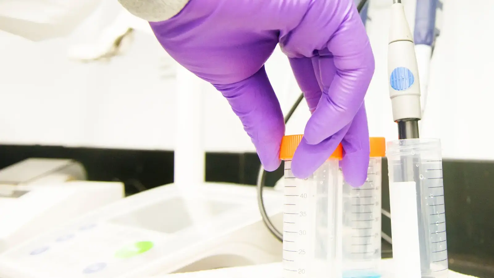 A person wearing purple gloves is holding a clear plastic test tube with an orange cap. The test tube is next to another similar tube and a pipette on a laboratory bench. Equipment is blurred in the background.