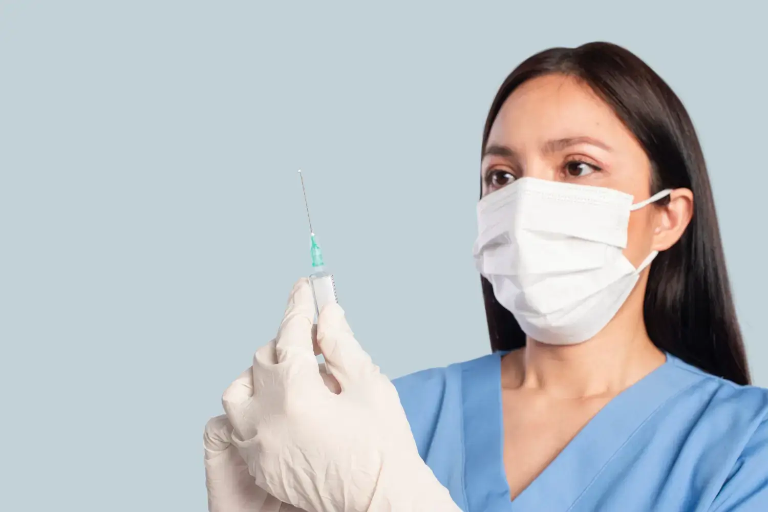 A healthcare professional in blue scrubs and a face mask holds a syringe, carefully inspecting it. The person is wearing white gloves and standing against a plain light gray background.