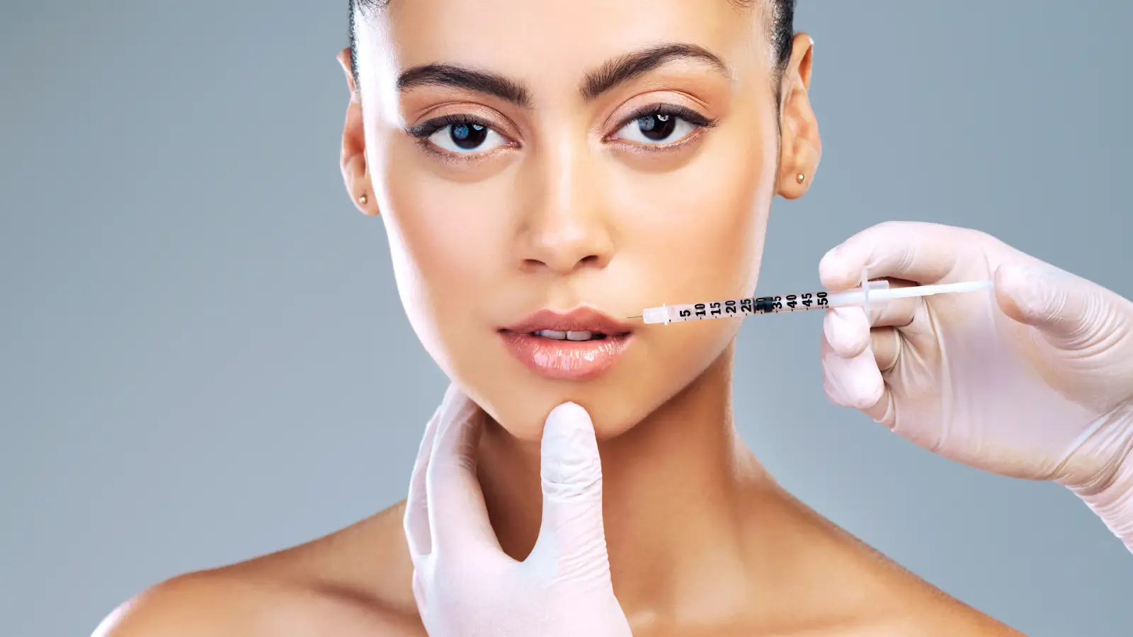 A woman with bare shoulders is looking straight ahead as a gloved hand holds a syringe near her lips.