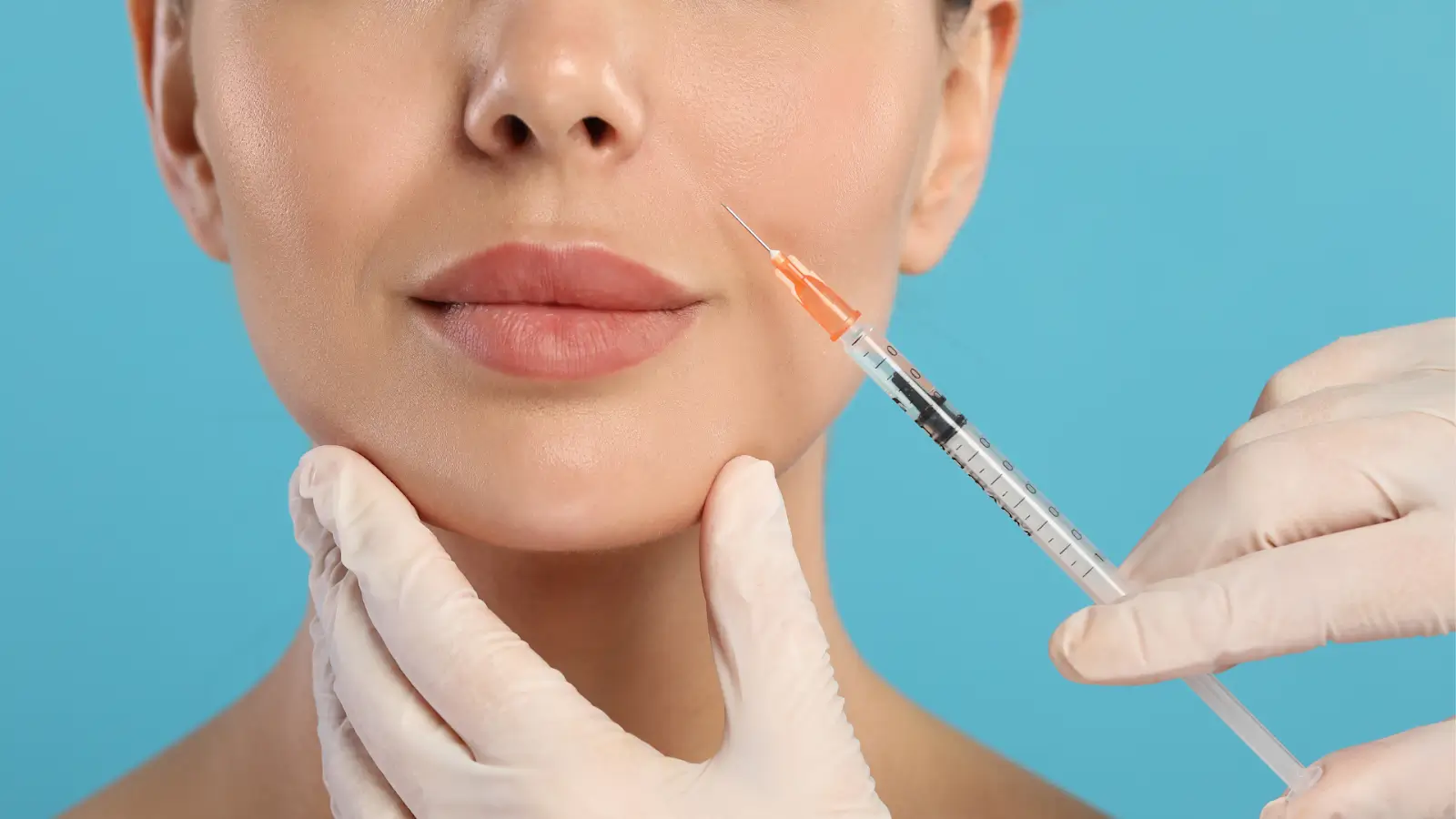 Close-up of a woman's lower face with plump lips, against a blue background. A gloved hand holds a syringe near her chin, possibly preparing for a cosmetic procedure.