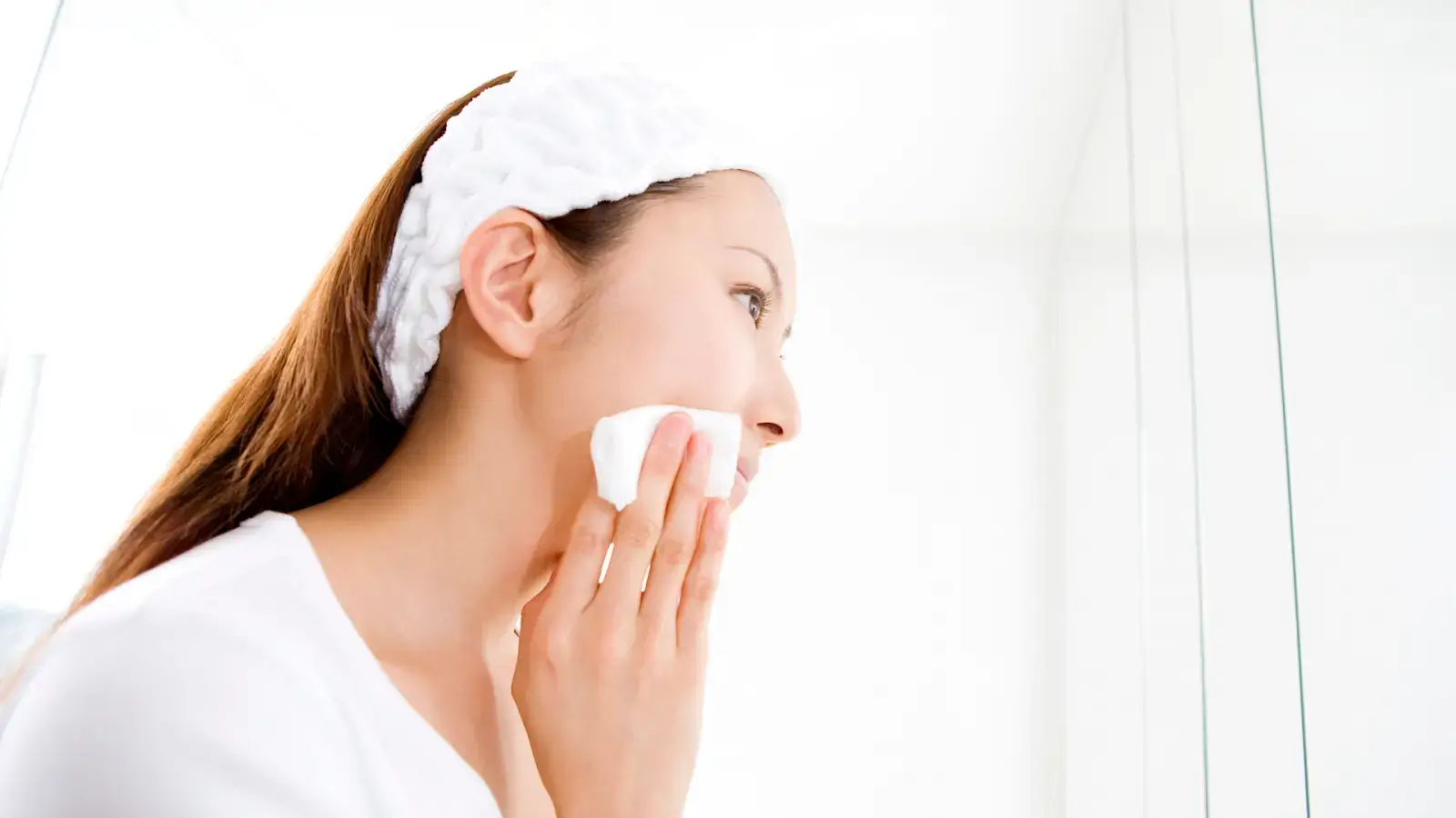 A woman with a white headband gently wipes her face with a cloth in a bright, minimalist setting. She appears focused on her skincare routine.