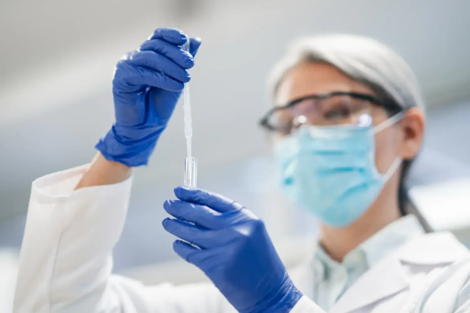 A scientist in a lab coat, safety glasses, and a mask uses a pipette to transfer liquid into a test tube. They are wearing blue gloves in a lab setting with a blurred background.