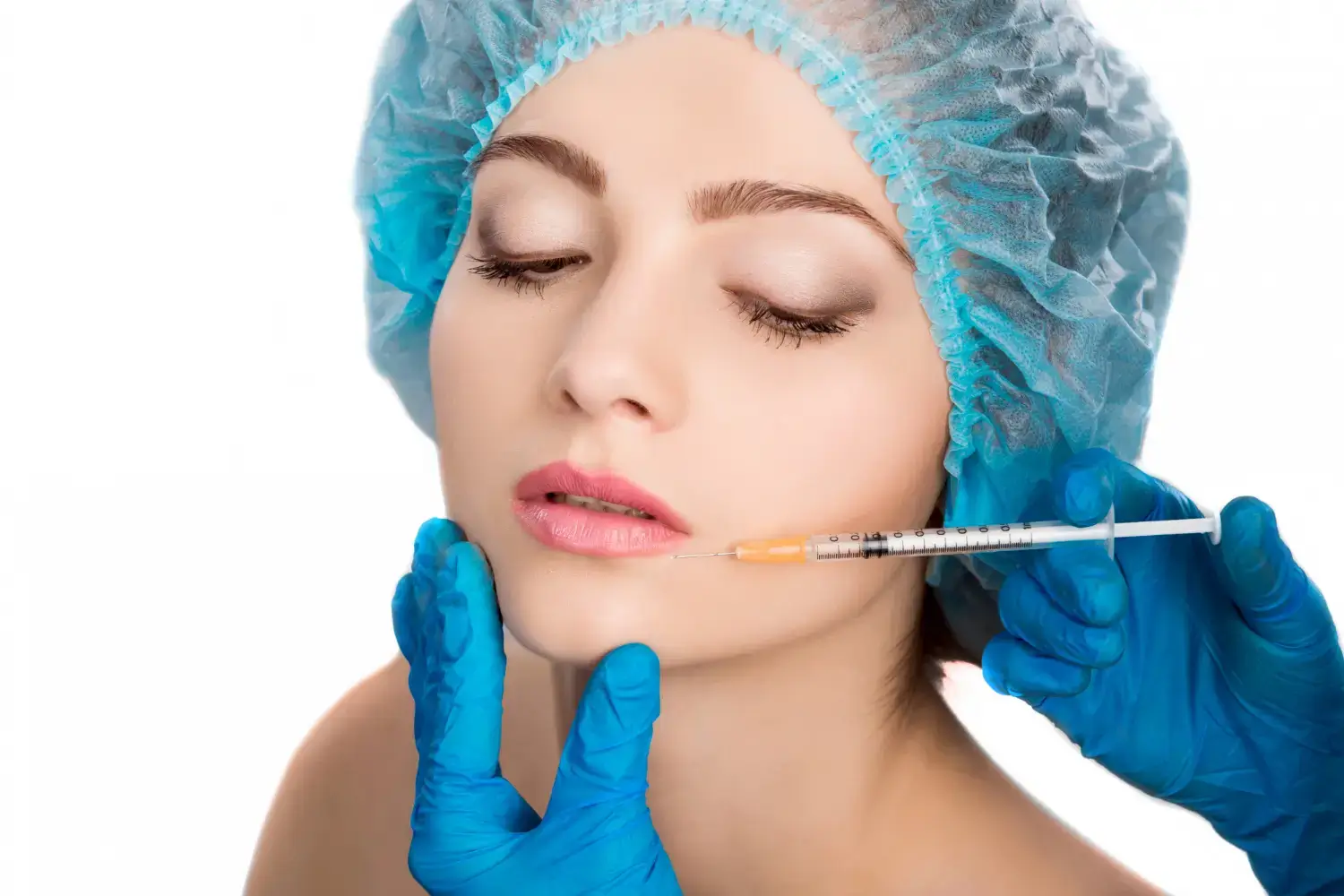 A young person with closed eyes is being administered an injection to the lips by hands in blue gloves. They are wearing a blue surgical cap. The background is white.