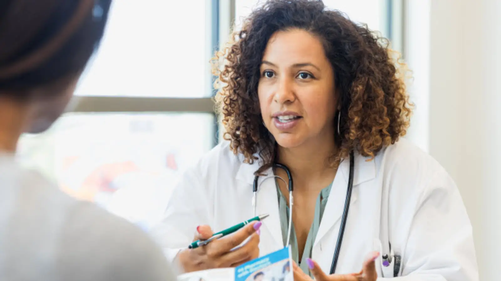 A doctor with curly hair converses with a patient, holding a pen and a document. She wears a white coat and has a stethoscope around her neck. They are seated at a table in a bright room with large windows in the background.