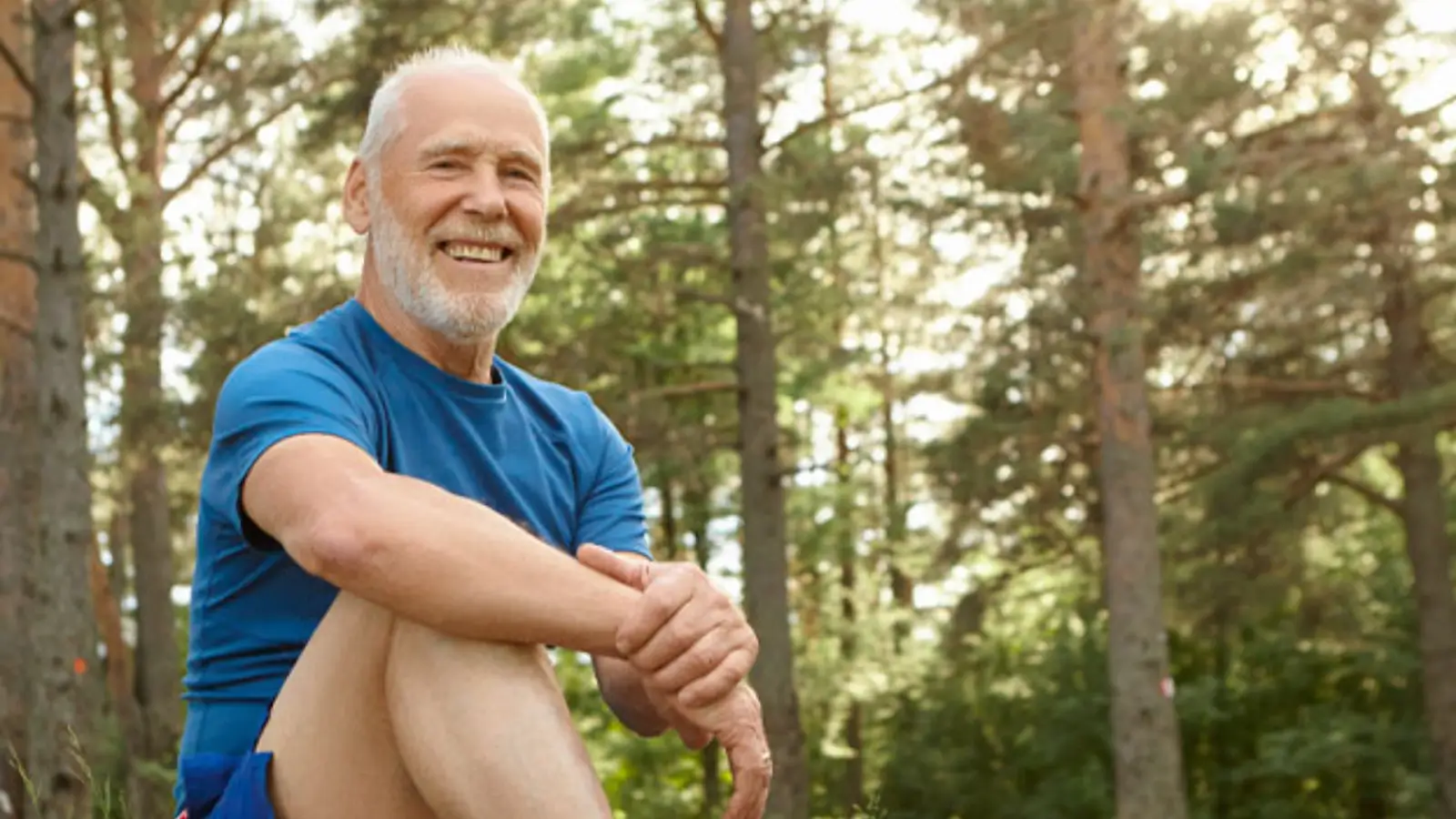 A smiling elderly man with a white beard sits cross-legged outdoors, wearing a blue shirt. He is surrounded by tall trees, with sunlight filtering through the leaves, creating a peaceful, natural setting.