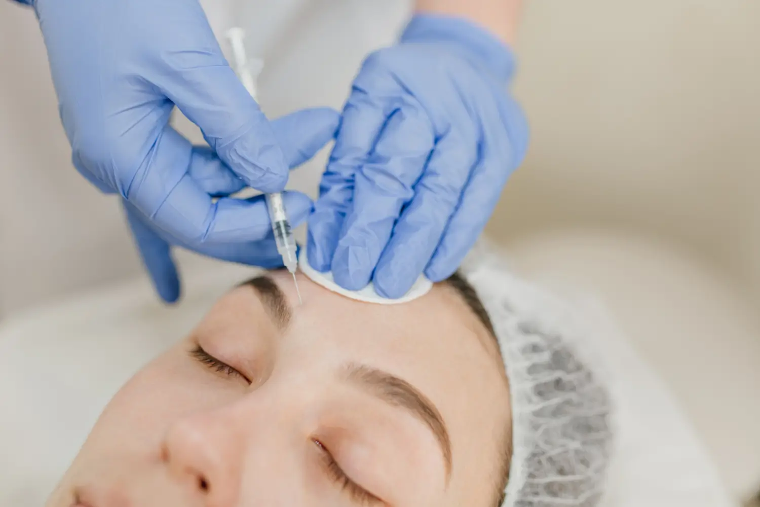 A person receives a cosmetic injection on their forehead. They are lying down with eyes closed, wearing a hair cover. 