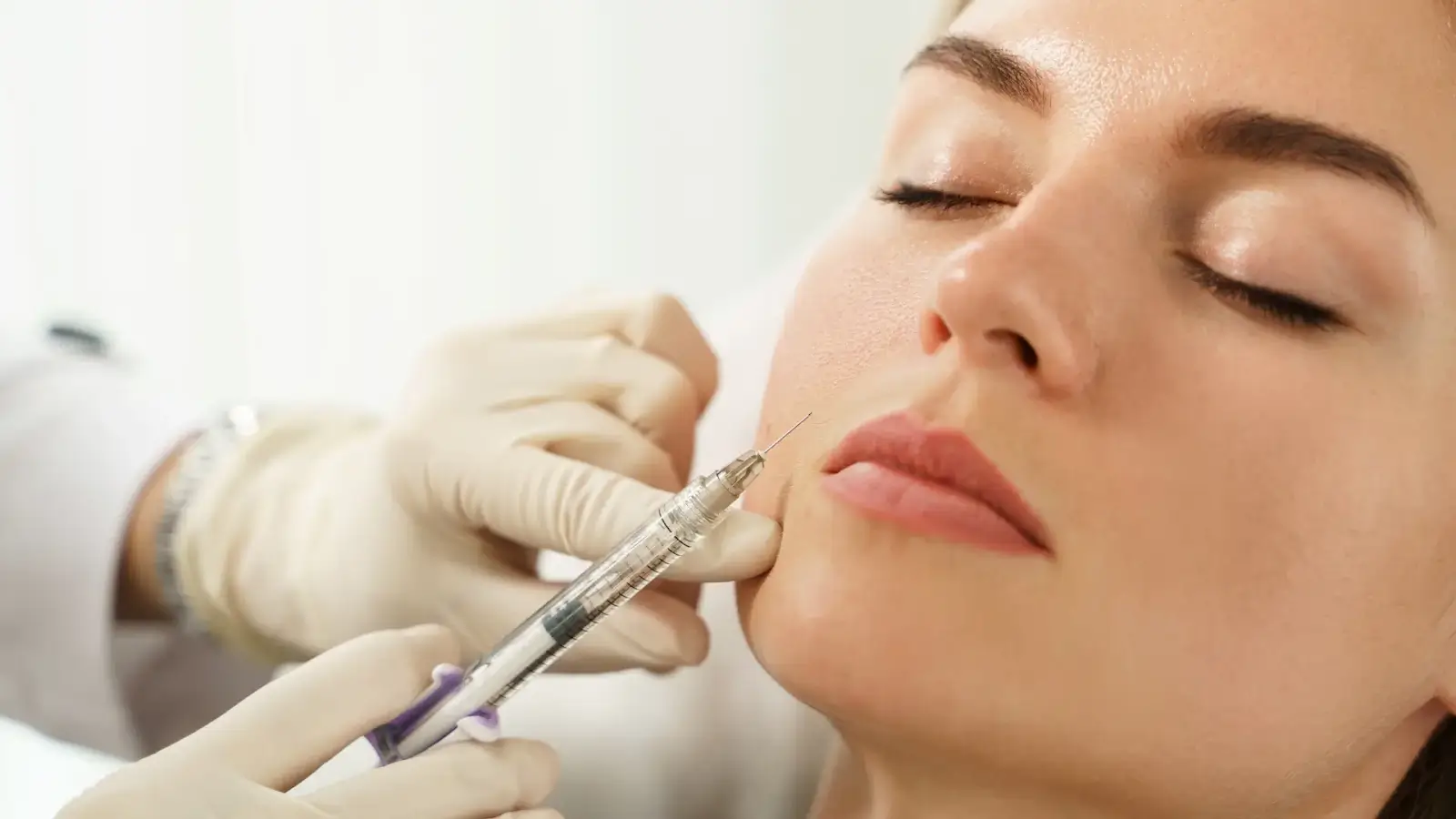 A person receiving a cosmetic injection in the face. A medical professional wearing white gloves is administering the treatment with a syringe. The person has closed eyes and relaxed expression, suggesting they are calm and at ease.