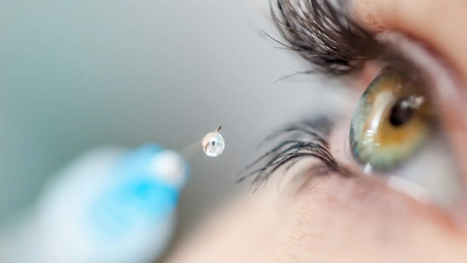 Close-up of an eye with a dropper releasing a liquid droplet near it. The eye is green with visible eyelashes. The background is blurred, highlighting the droplet and eye.