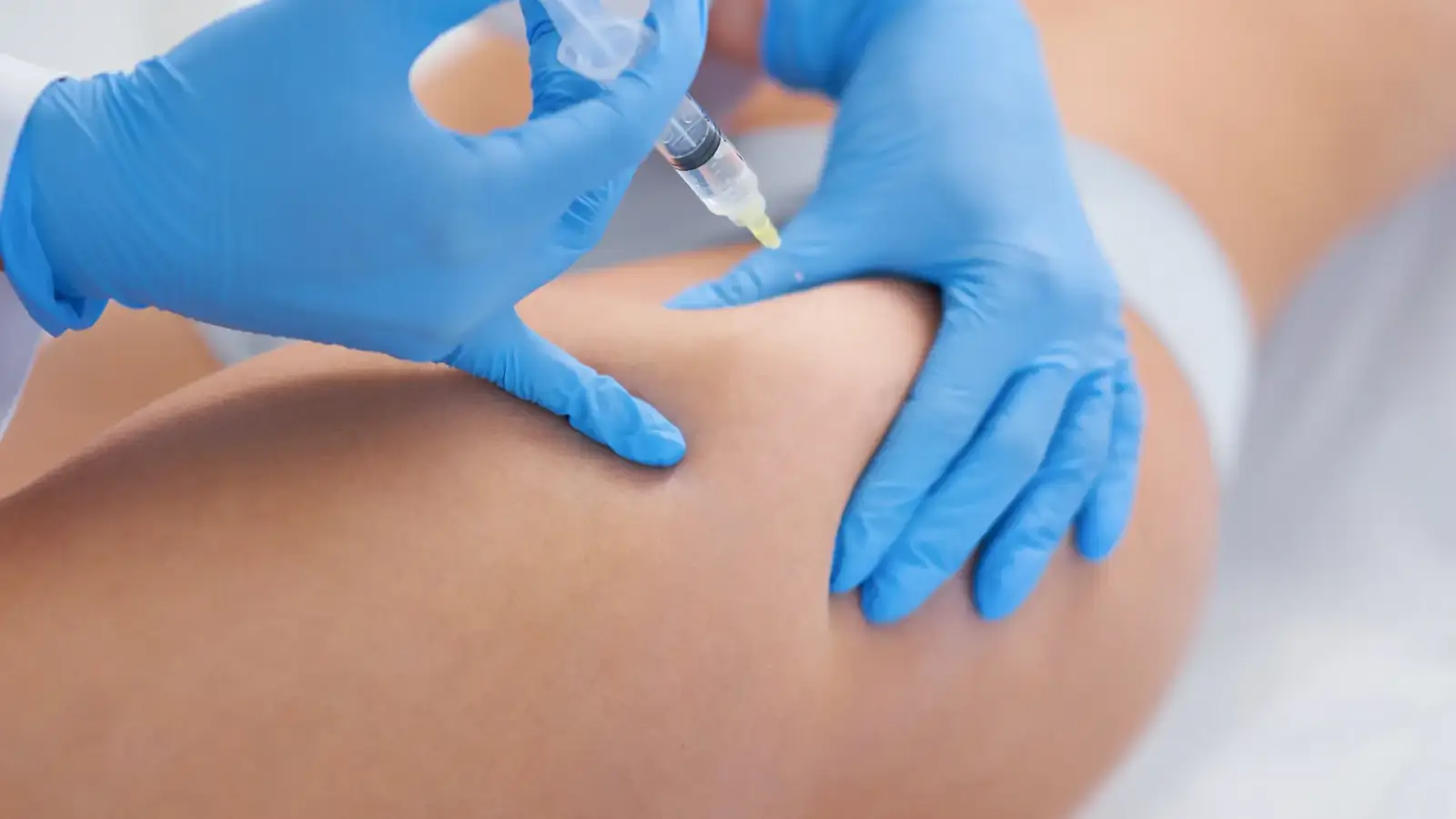 A medical professional in blue gloves injects a syringe into a person's thigh. The background is neutral and out of focus.