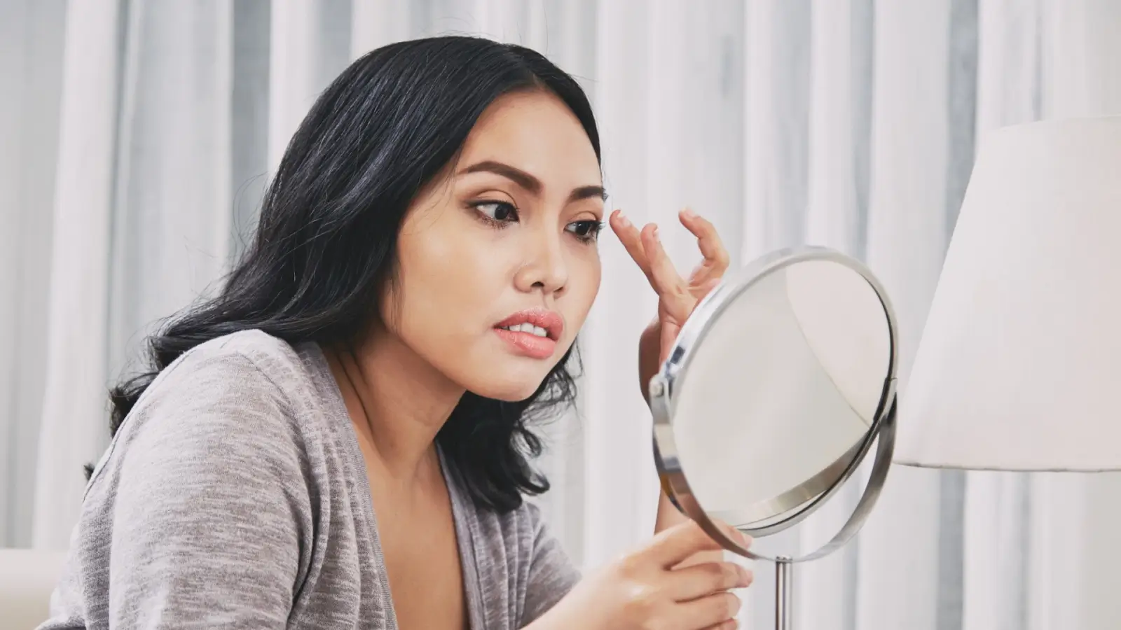 A person with long black hair is intently looking into a round mirror while applying makeup to their eyelid. They are wearing a gray top, and the background features soft, white curtains.