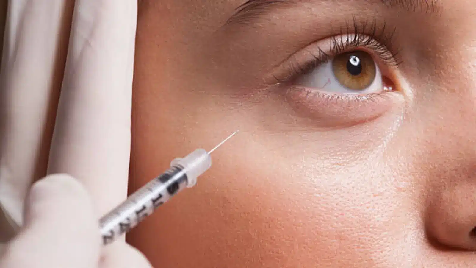 Close-up of a person receiving an injection near the eye. A gloved hand holds a syringe against the skin below the eye, with the needle poised for injection. The person's eye is wide open, showing details of their eyelashes and iris.