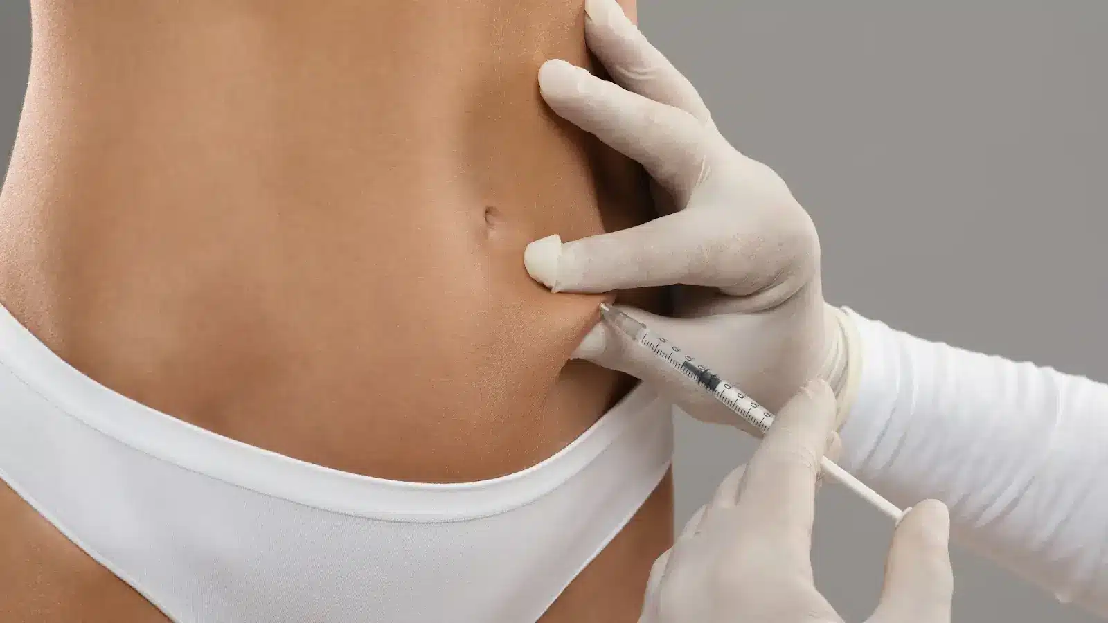 A person wearing white underwear is receiving an injection in their abdomen. A gloved hand is administering the shot with a syringe. The background is plain and gray.