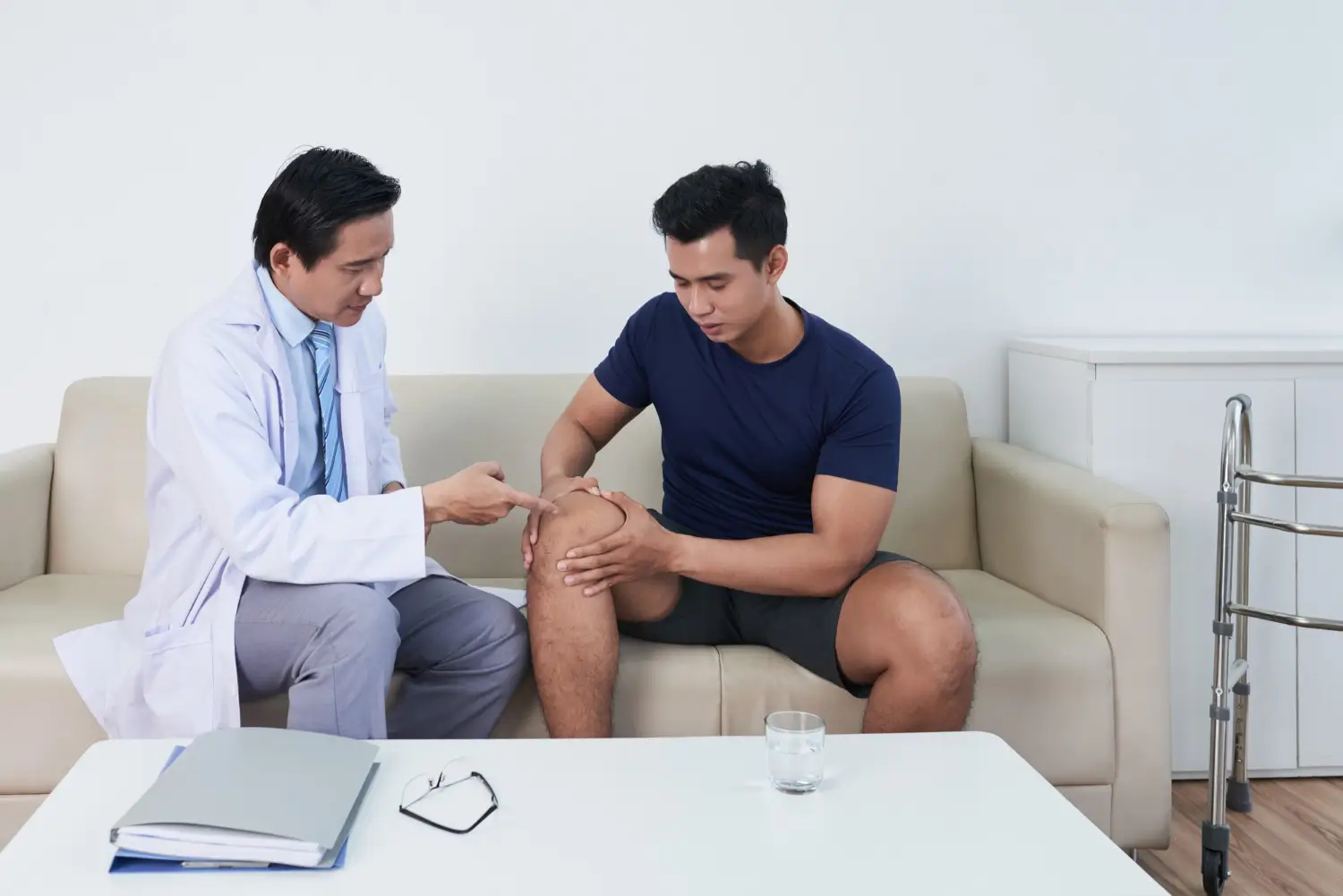 A doctor in a white coat examines a patient's knee as they sit on a sofa in a medical office.