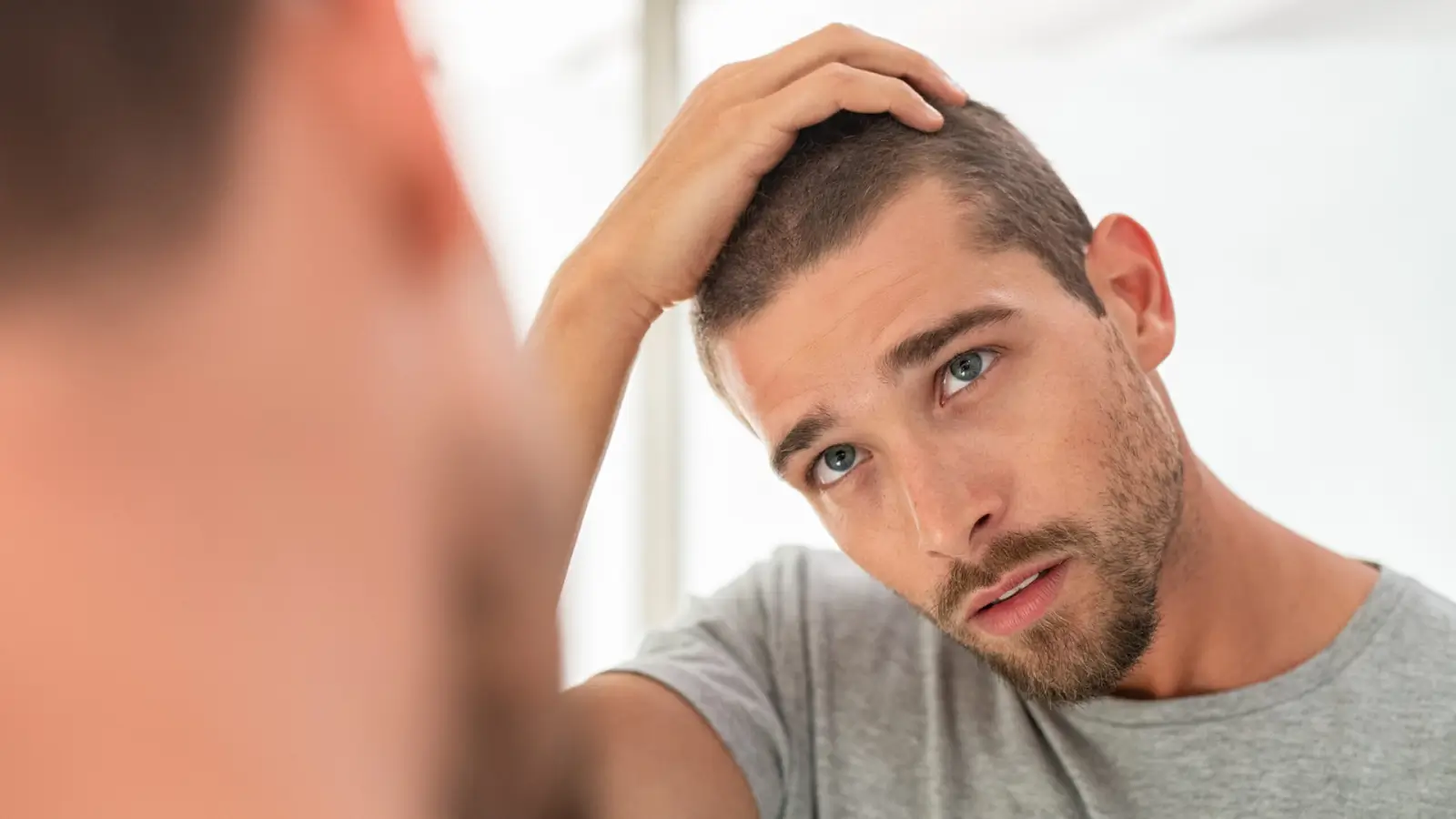 A man with a short beard and short hair looks at himself in the mirror, touching his head. He is wearing a gray T-shirt and appears to be examining his hair.