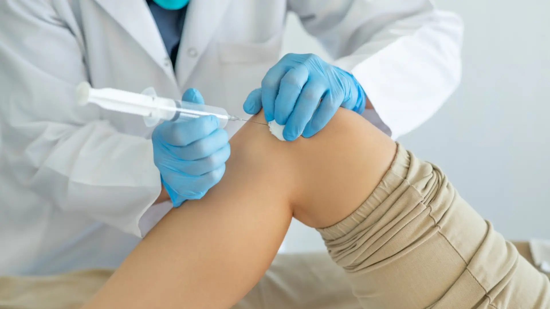 A medical professional in a white coat and blue gloves administers an injection to a patient's knee. 