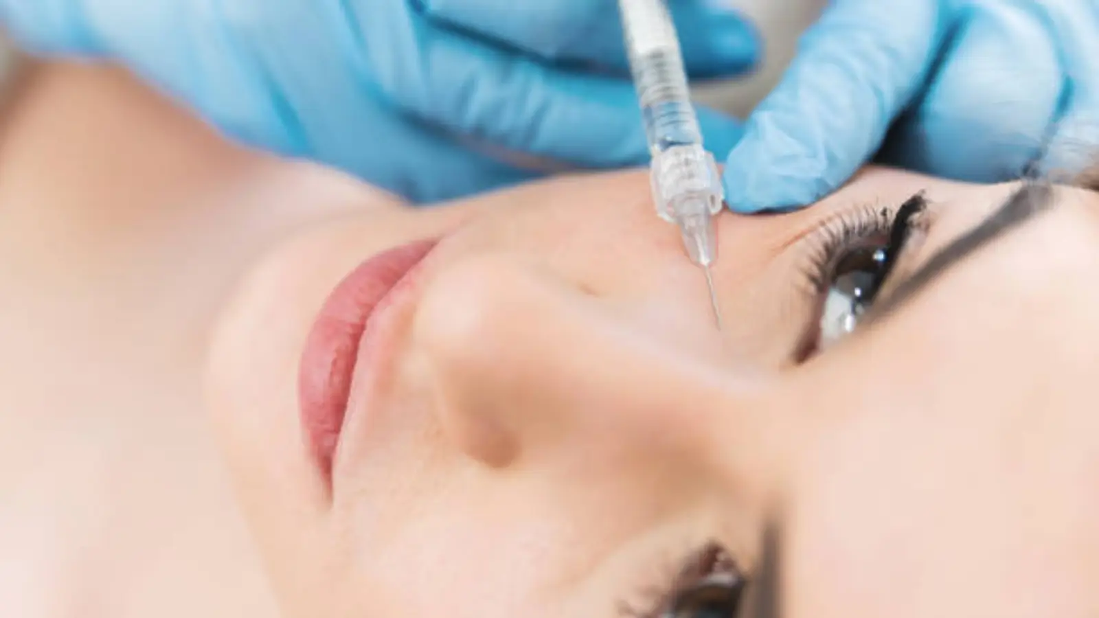 A close-up of a person receiving a cosmetic injection near the eye. A gloved hand holds a syringe, and the person's face is tilted upward with eyes open, focusing on the procedure.