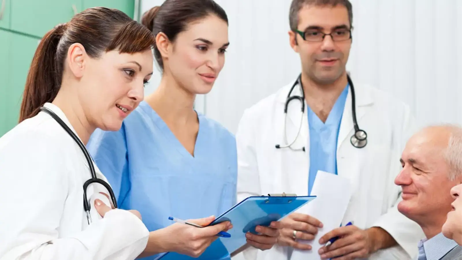 A group of healthcare professionals, including two women and two men, are in discussion. The women are holding a clipboard and wearing scrubs, while the men wear white coats with stethoscopes. A smiling older man is seated nearby.