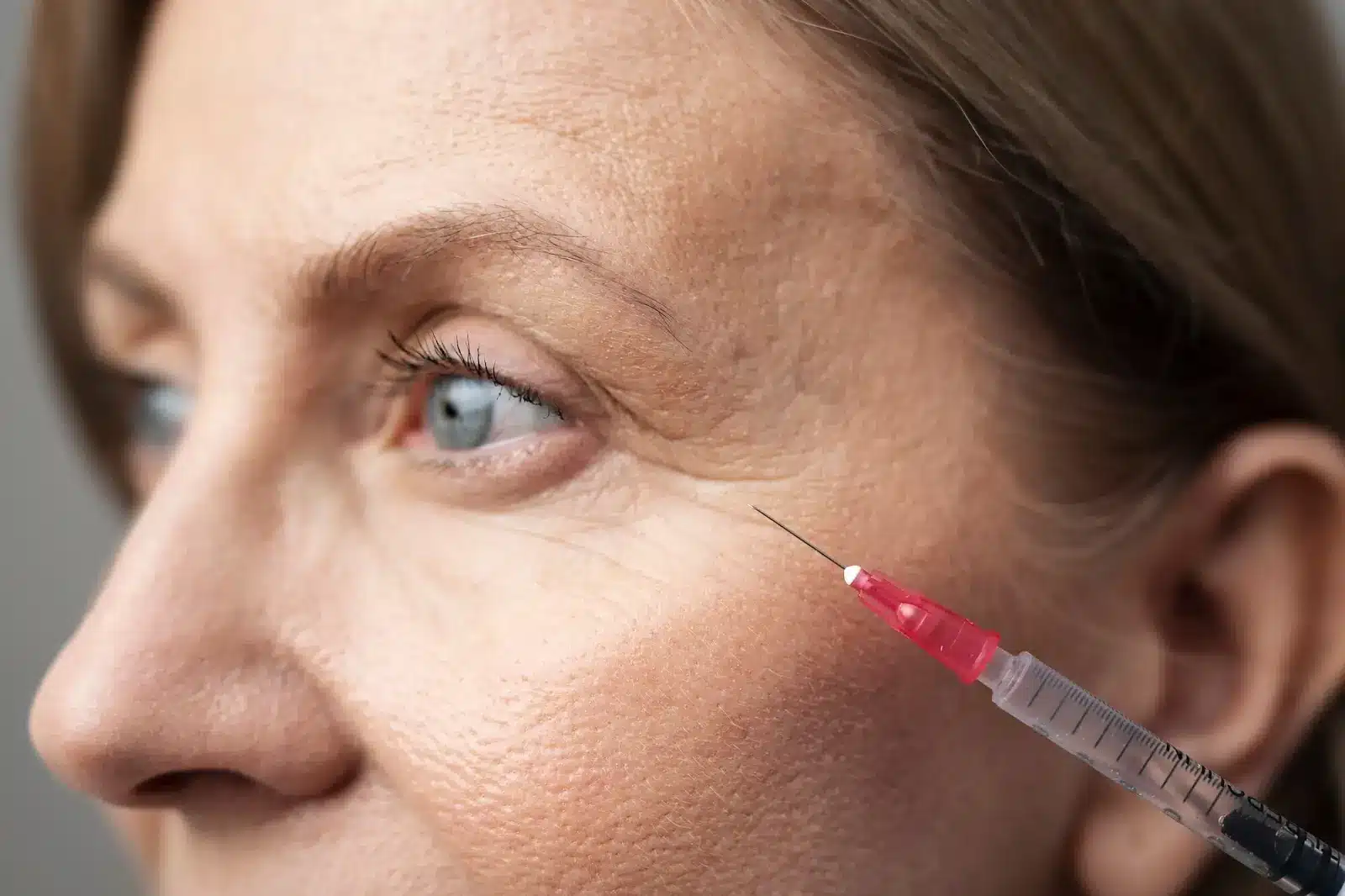 Close-up of a woman's face with a focus on an eye. A syringe with a red-tinted tip is positioned near the outer corner of her eye, suggesting a cosmetic procedure. The skin appears textured and natural.