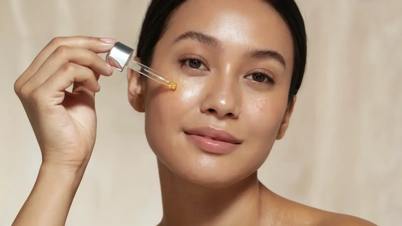 A person smiling and applying facial serum with a dropper to their cheek. The background is a soft, neutral color, enhancing the serene and skincare-focused atmosphere.