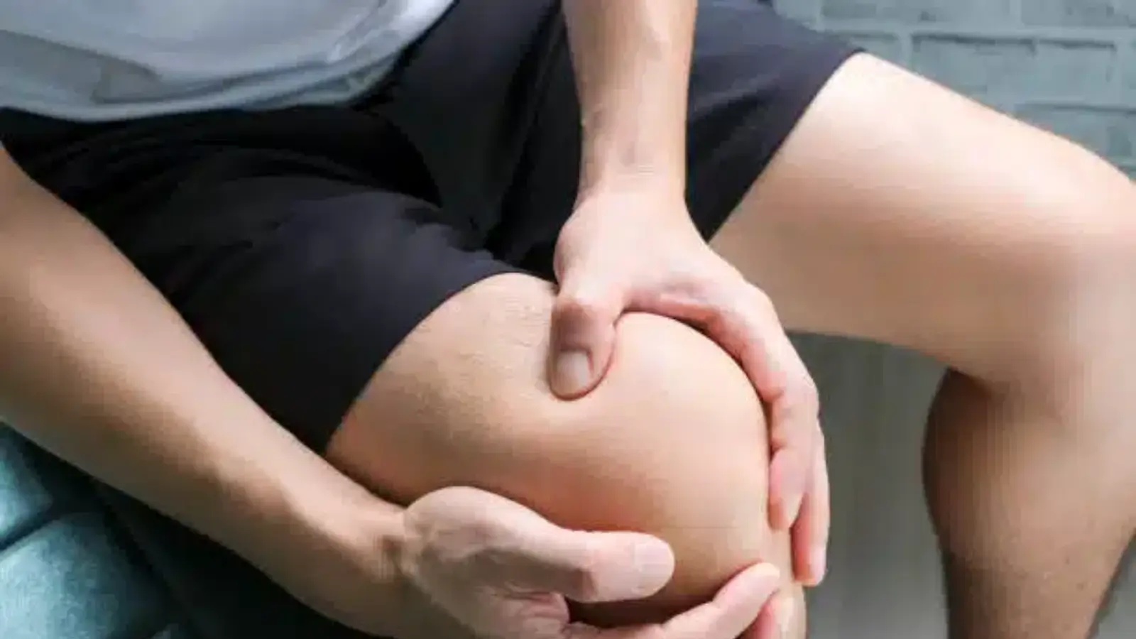 A person wearing black shorts is sitting and holding their right knee with both hands, indicating discomfort or pain. The focus is on the knee and hands.