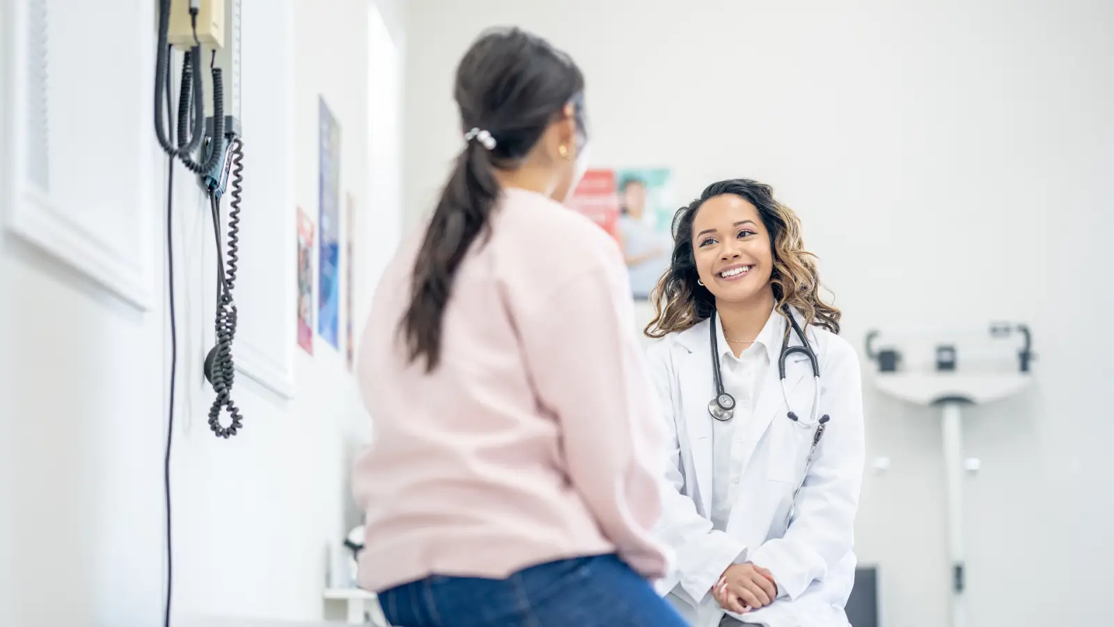 A healthcare professional in a white coat, wearing a stethoscope, is smiling while talking to a seated patient in casual attire. They are in a medical examination room with various medical equipment visible in the background.