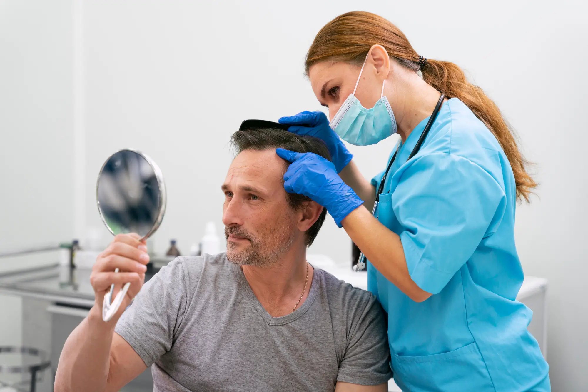 A man with graying hair examines his hair in a handheld mirror while a healthcare professional in a blue scrub top and face mask inspects his scalp.