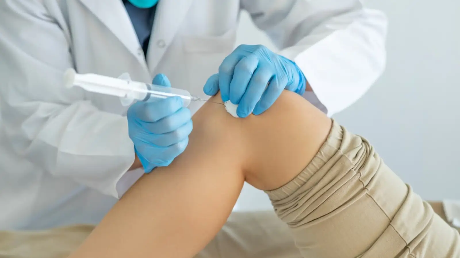 A person in a white coat and blue gloves administers an injection into a patient's bent knee. The patient is wearing khaki shorts. The image suggests a medical procedure or treatment.