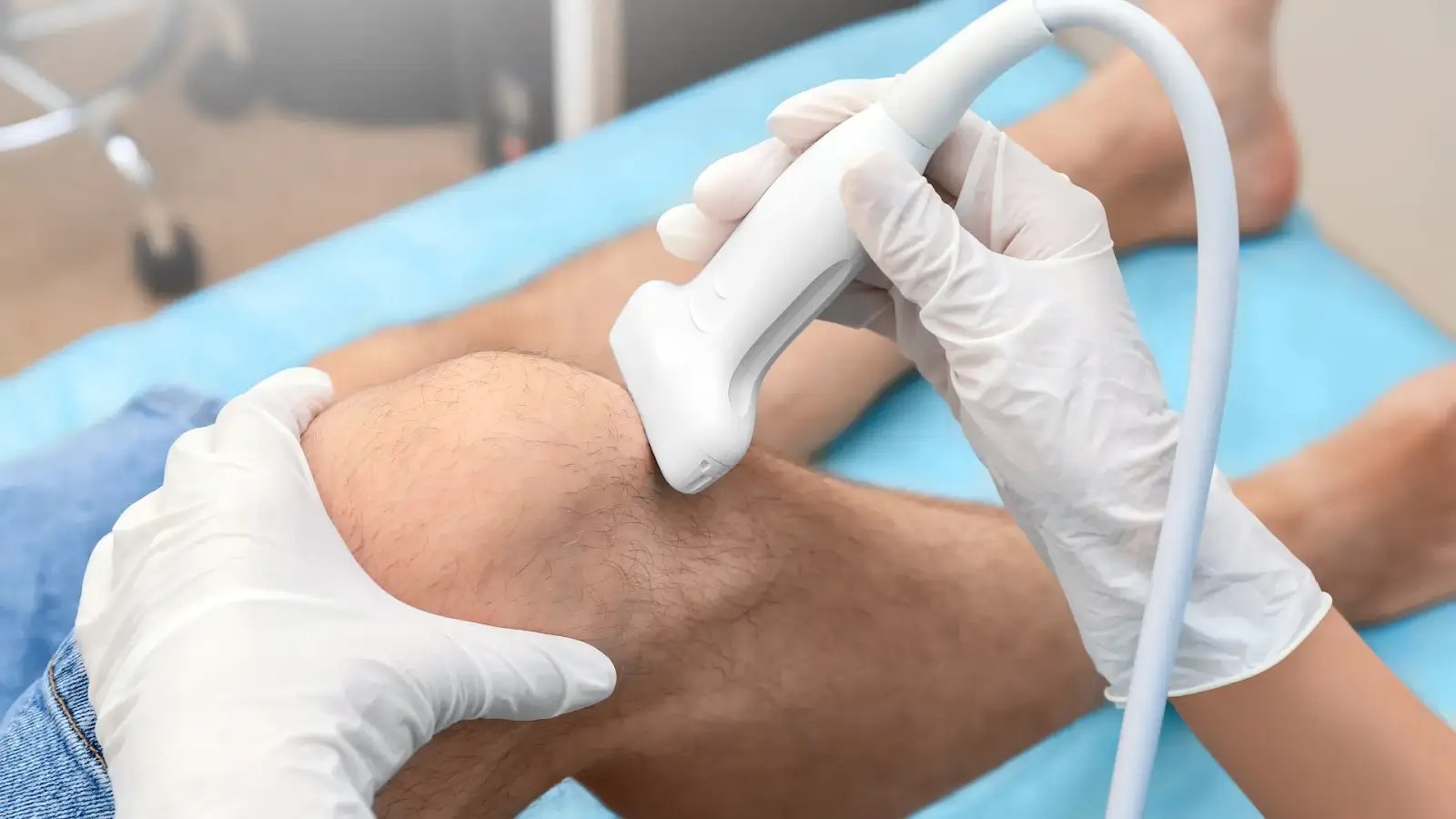 A medical professional wearing gloves is using an ultrasound device on a patient's knee. The patient is lying down on a blue sheet, with the leg partially covered by shorts. The scene suggests a non-invasive diagnostic procedure.