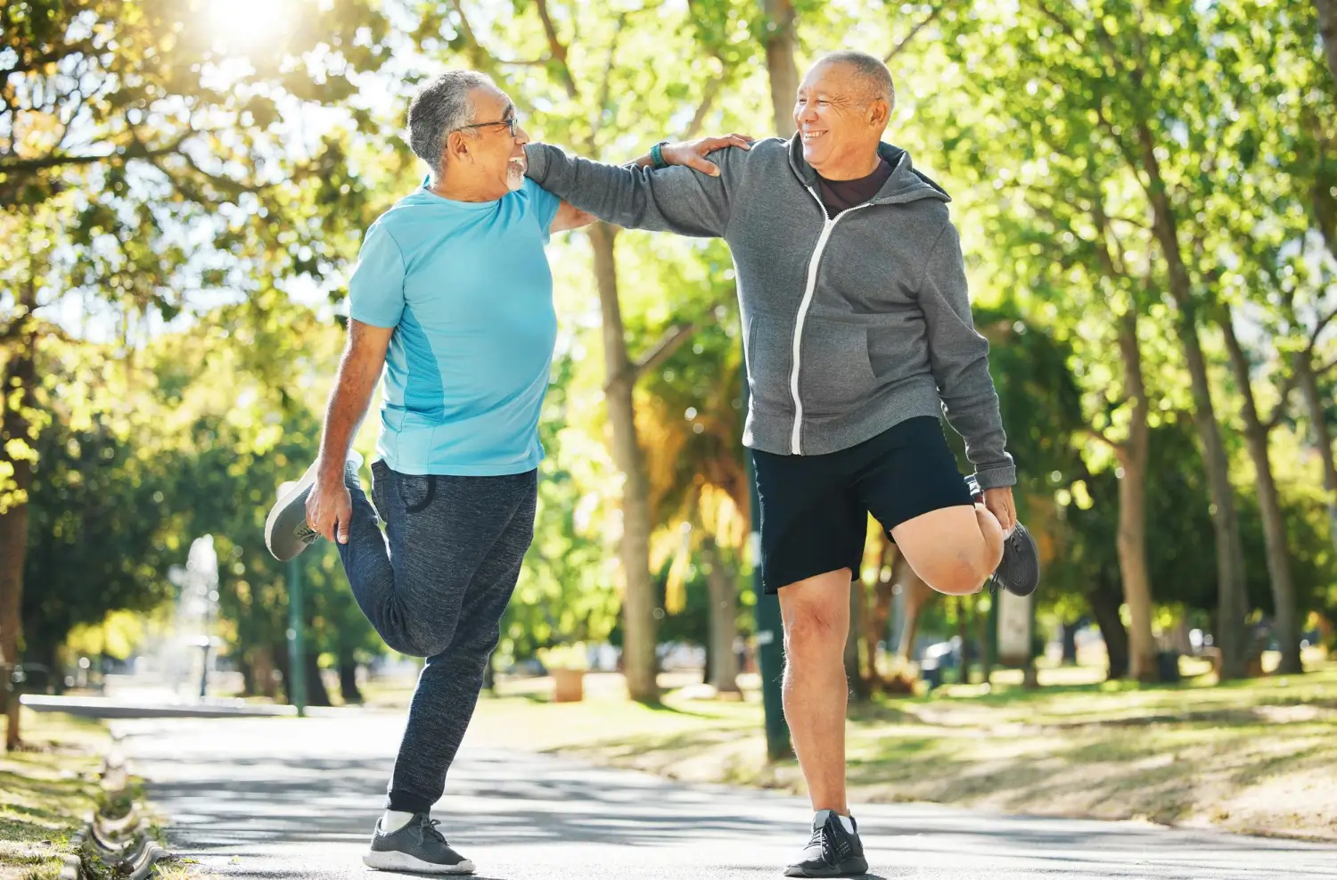 Two elderly men stretch their legs in a sunlit park. They are smiling and chatting, dressed in casual athletic wear.