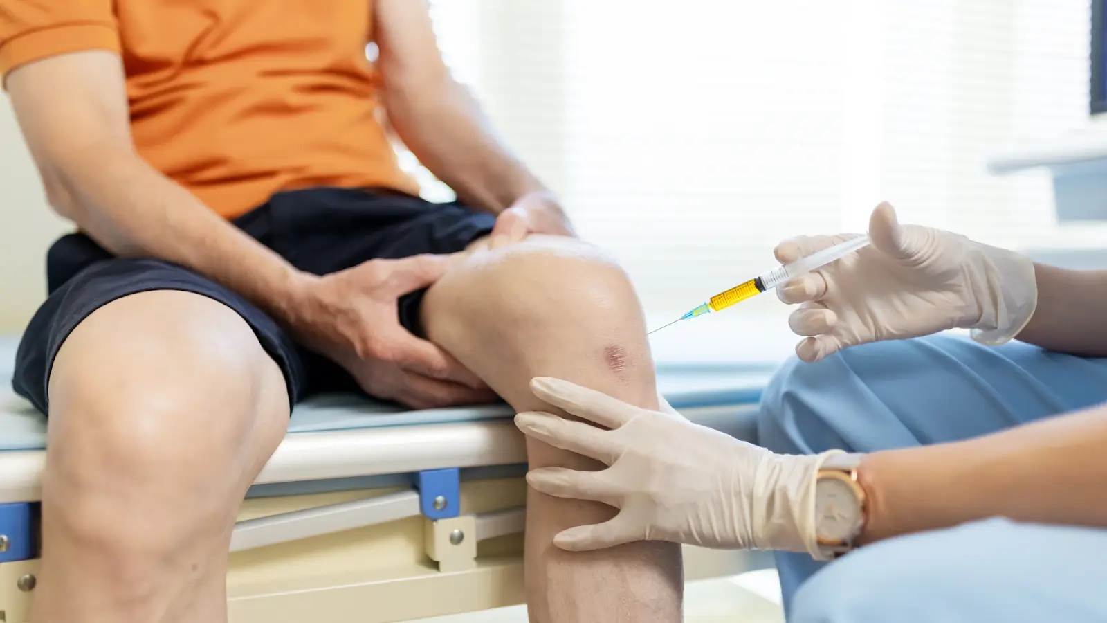 A person sitting on a medical examination table receives an injection in the knee from a healthcare professional wearing gloves.