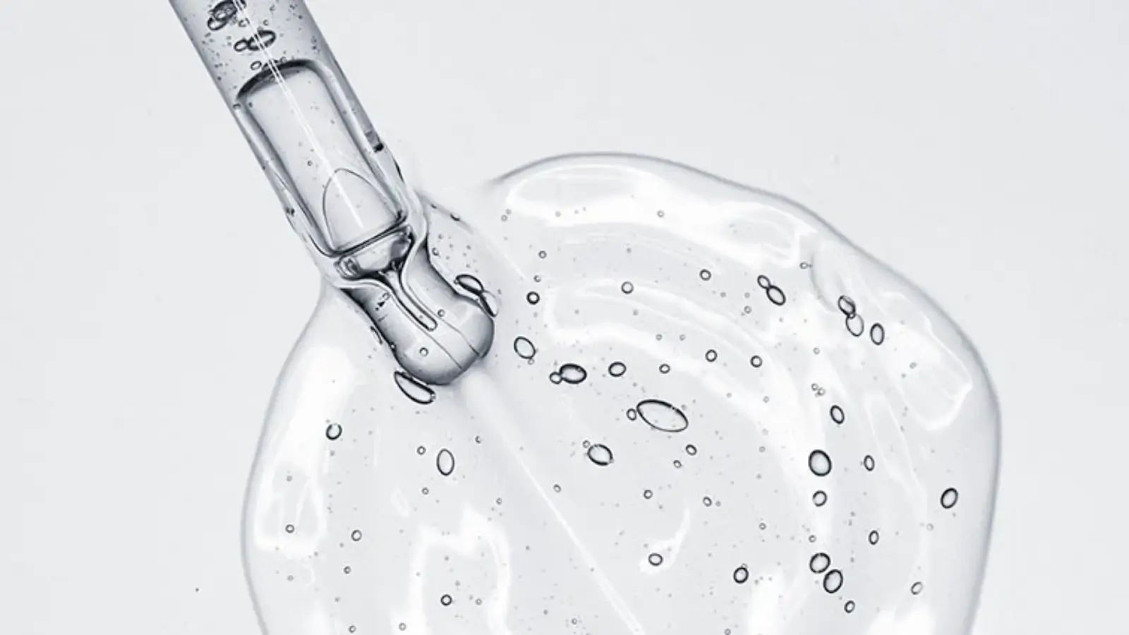 Close-up of a clear liquid gel being dispensed from a dropper. The gel is viscous with small air bubbles visible, creating a textured surface beneath the dropper tip. The background is plain white, highlighting the transparency and texture of the gel.