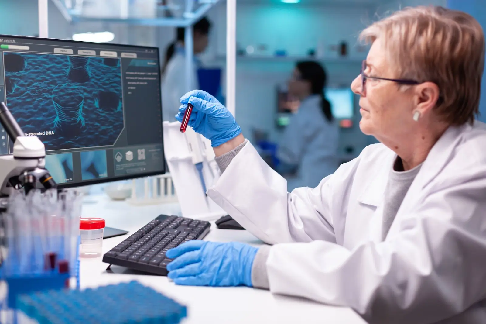 A scientist in a lab coat and blue gloves examines a test tube with a red liquid while seated at a desk with a keyboard and a monitor displaying a magnified image of DNA. Lab equipment and another person are visible in the background.