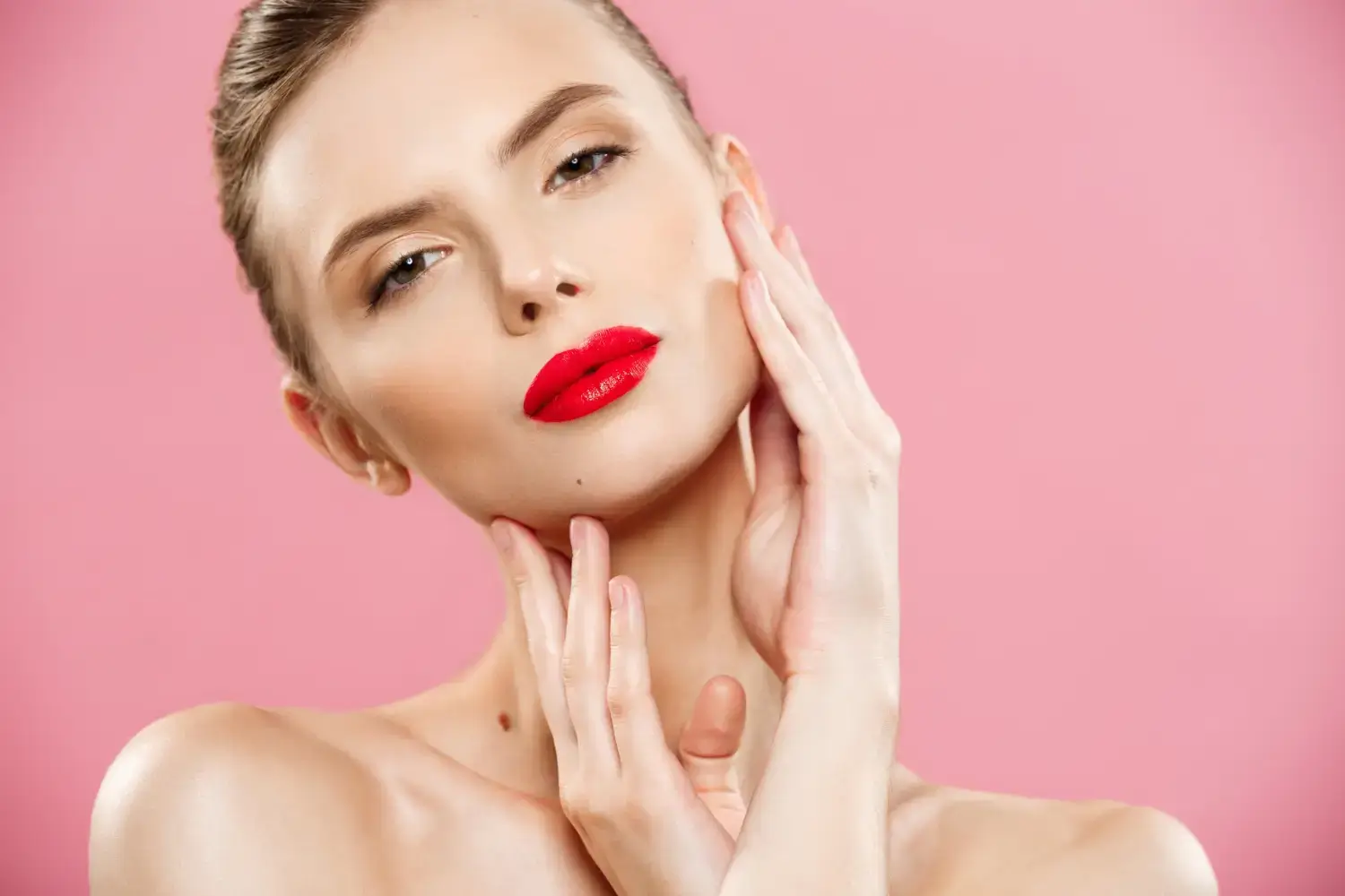 A person with red lipstick poses against a pink background, gently touching their face with both hands. They have a calm expression and sleek, pulled-back hair.