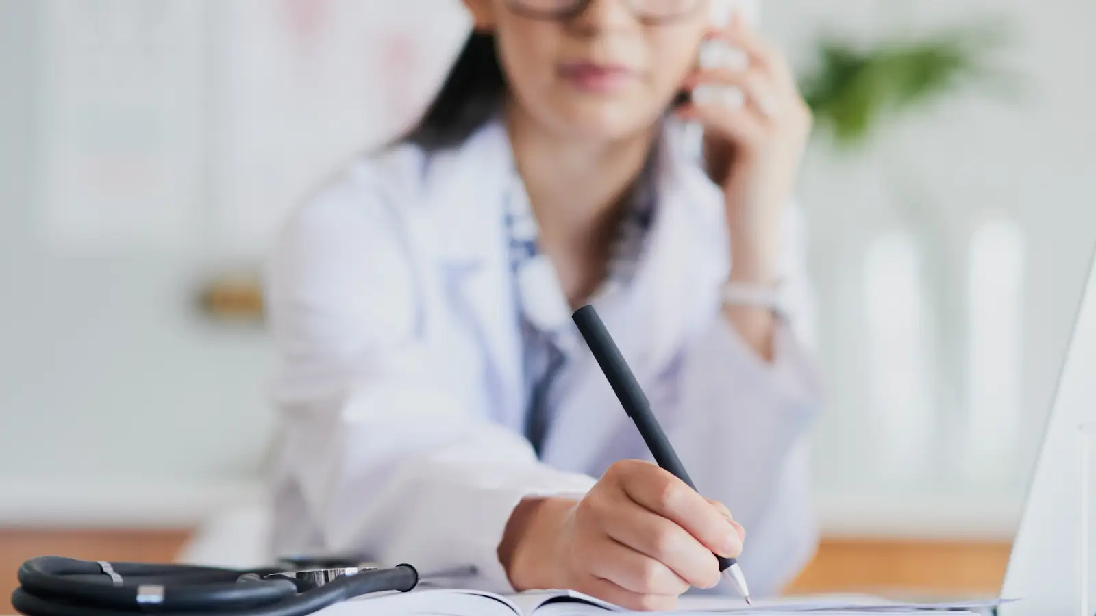 A person in a white coat is writing in a notebook on a desk, with a stethoscope nearby. Their face is blurred, focusing on their hand and pen. 