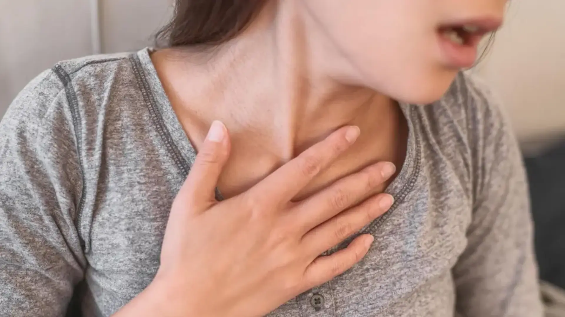 A person in a gray shirt holds their hand to their throat, appearing to be in discomfort.