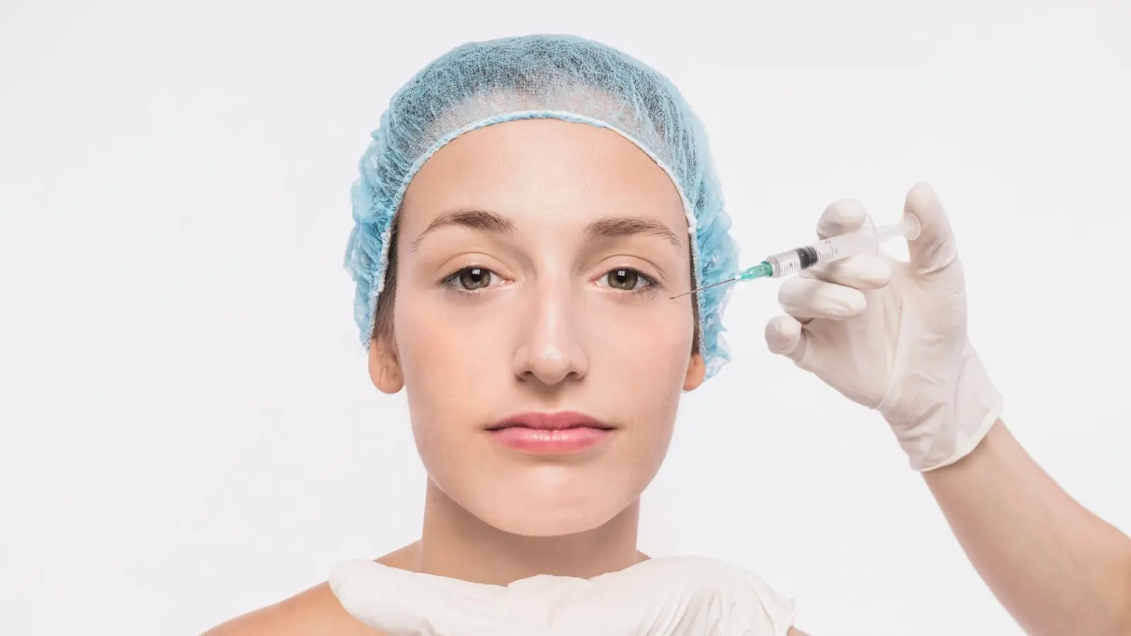 A person wearing a blue hair cap receives a facial injection from a gloved hand. The background is plain white. The person's expression is calm.