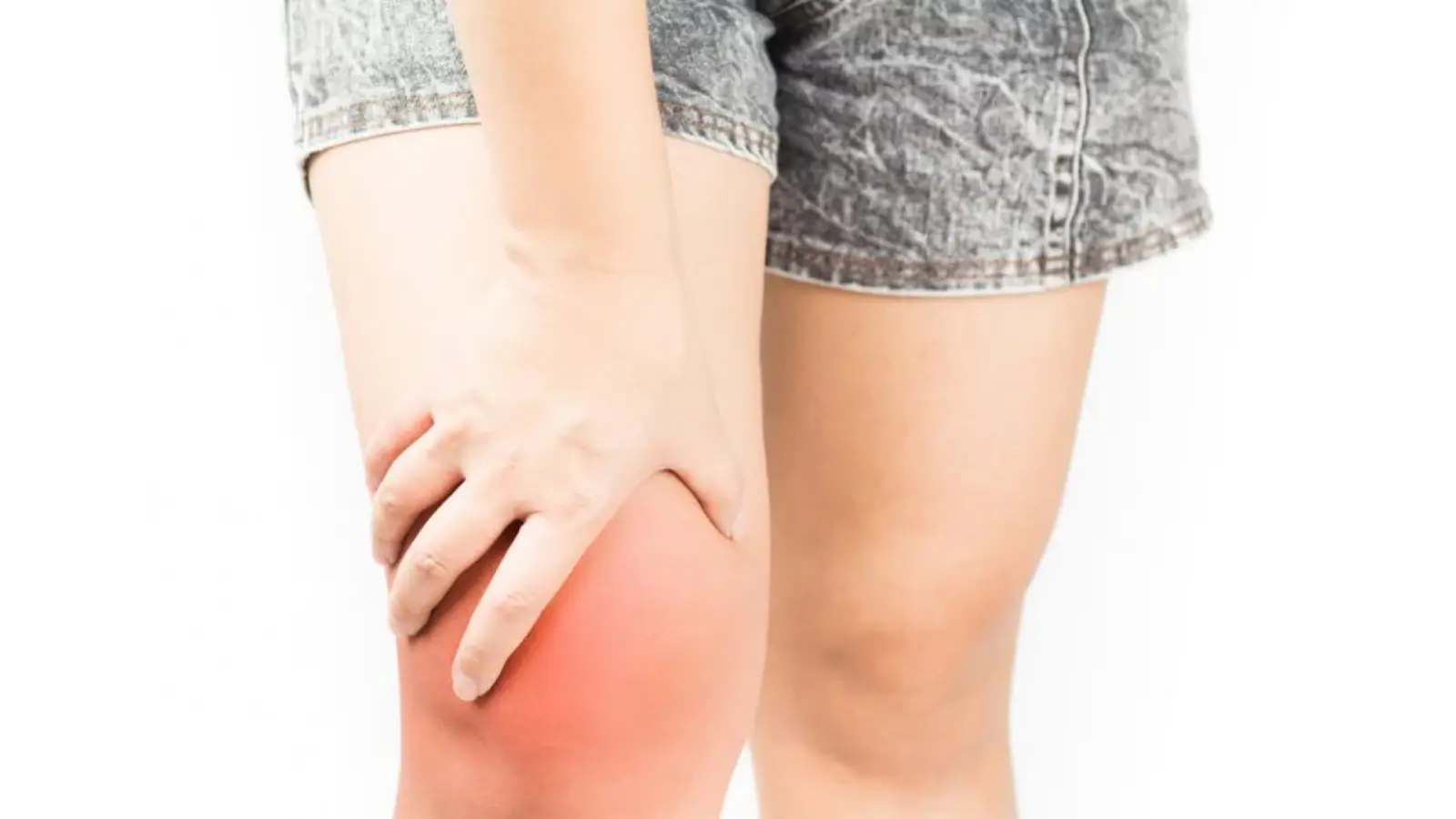 Person wearing denim shorts holding a swollen, red knee with one hand, indicating pain or discomfort.