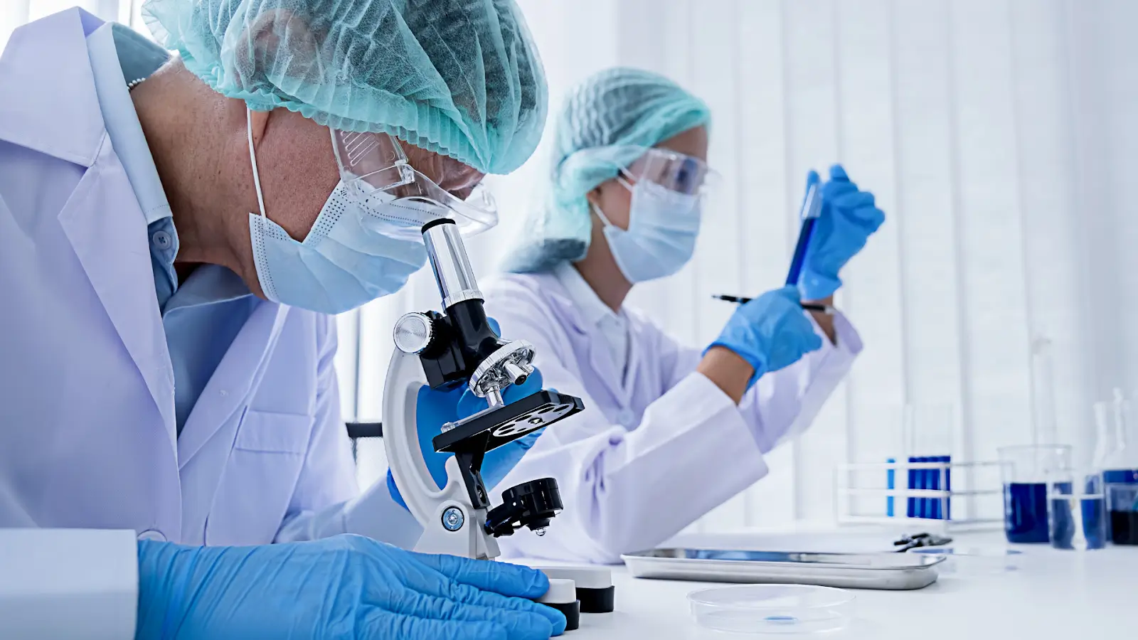 Two scientists in protective gear work in a lab. One peers into a microscope, while the other examines a test tube. Both wear gloves, masks, and hair covers, surrounded by lab equipment on a white counter.