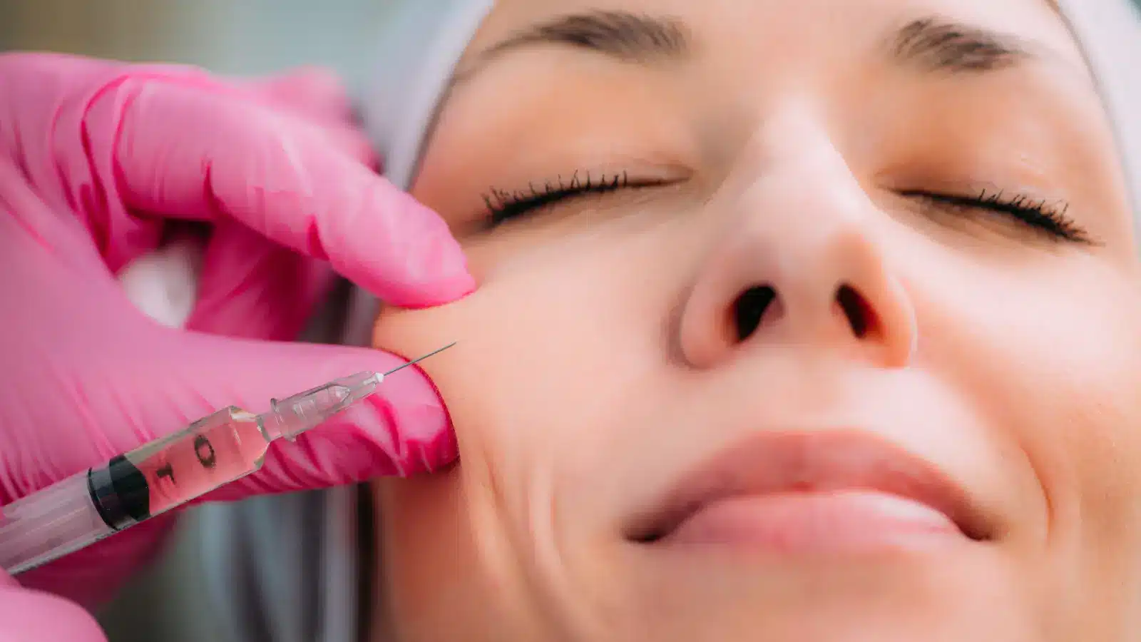 A person with closed eyes is receiving a facial injection. The injector is wearing pink gloves and holding a syringe near the person's cheek.