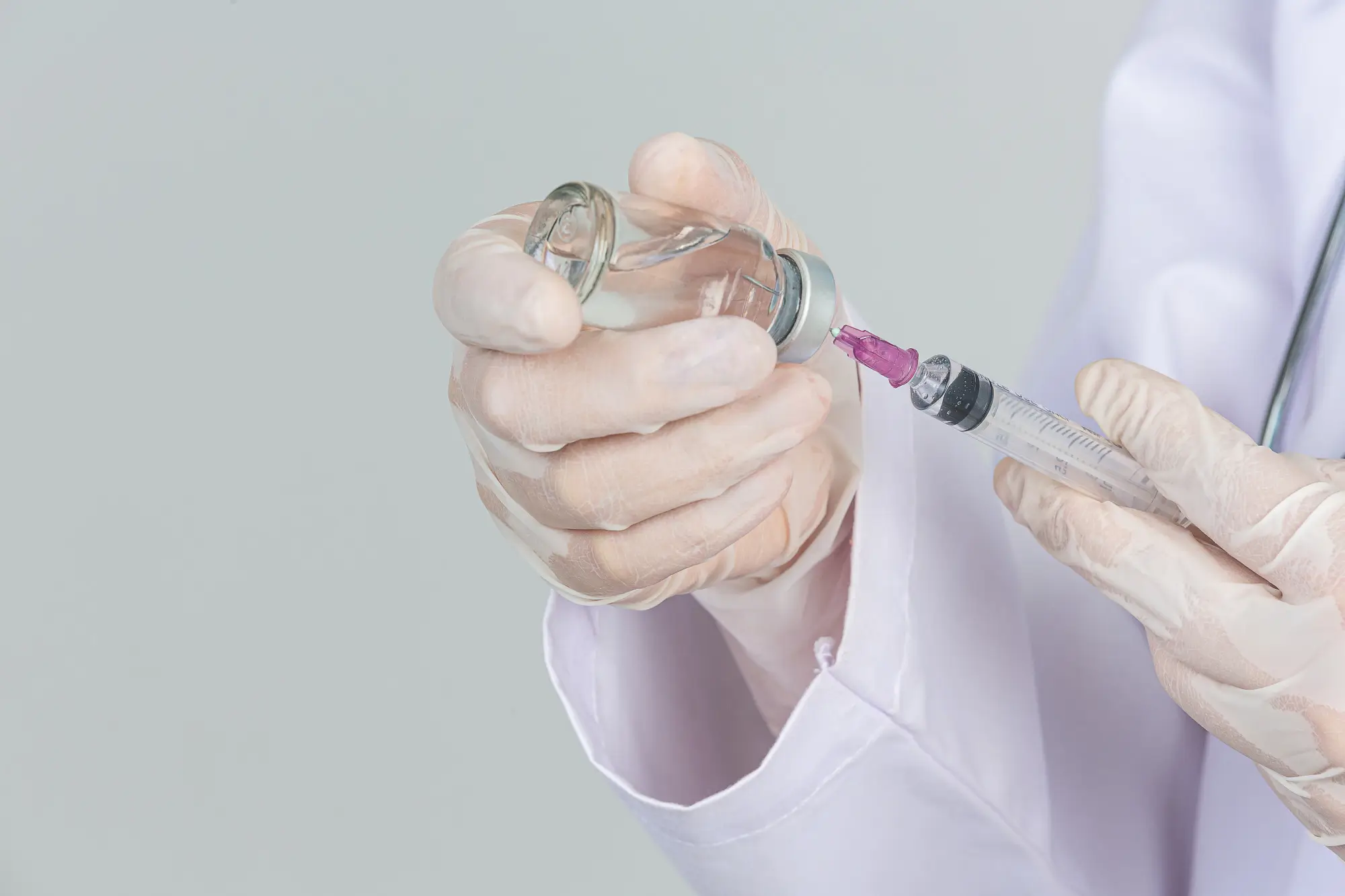 A person wearing gloves holds a vial and a syringe, drawing liquid from the vial. The background is a plain, light color, and the individual is dressed in a white lab coat.