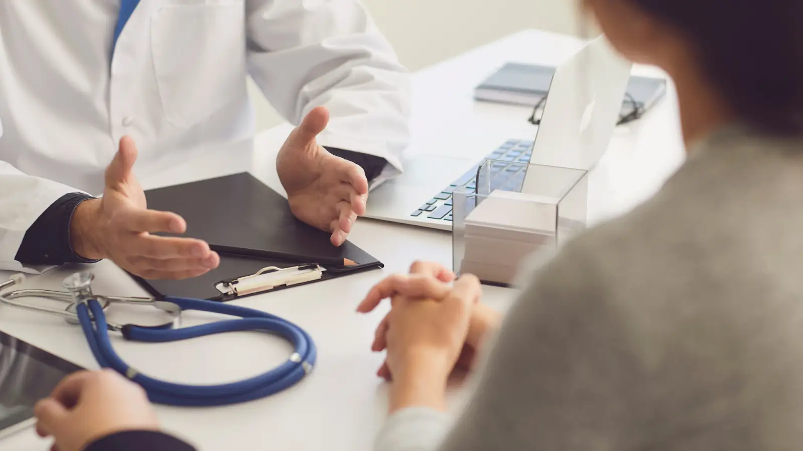 A doctor in a white coat is talking with a patient at a desk. The doctor's hands are gesturing, and a blue stethoscope, clipboard, laptop, and tissue box are on the table. The patient sits across, listening attentively with hands clasped.