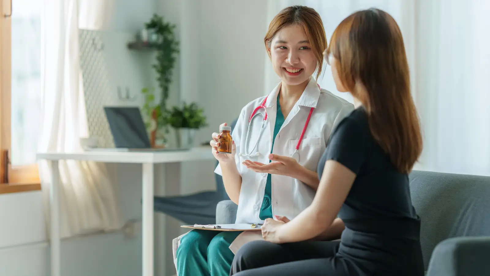 A healthcare professional with a stethoscope and clipboard converses with a seated woman, holding a medicine bottle. They are in a bright room with a desk, potted plant, and laptop in the background.