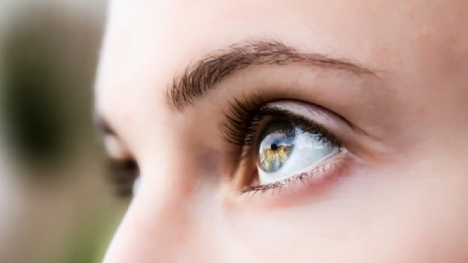 Close-up of a person's right eye gazing upward. The eye is light-colored with long eyelashes, and the eyebrow is partially visible. The background is softly blurred with green and brown tones.