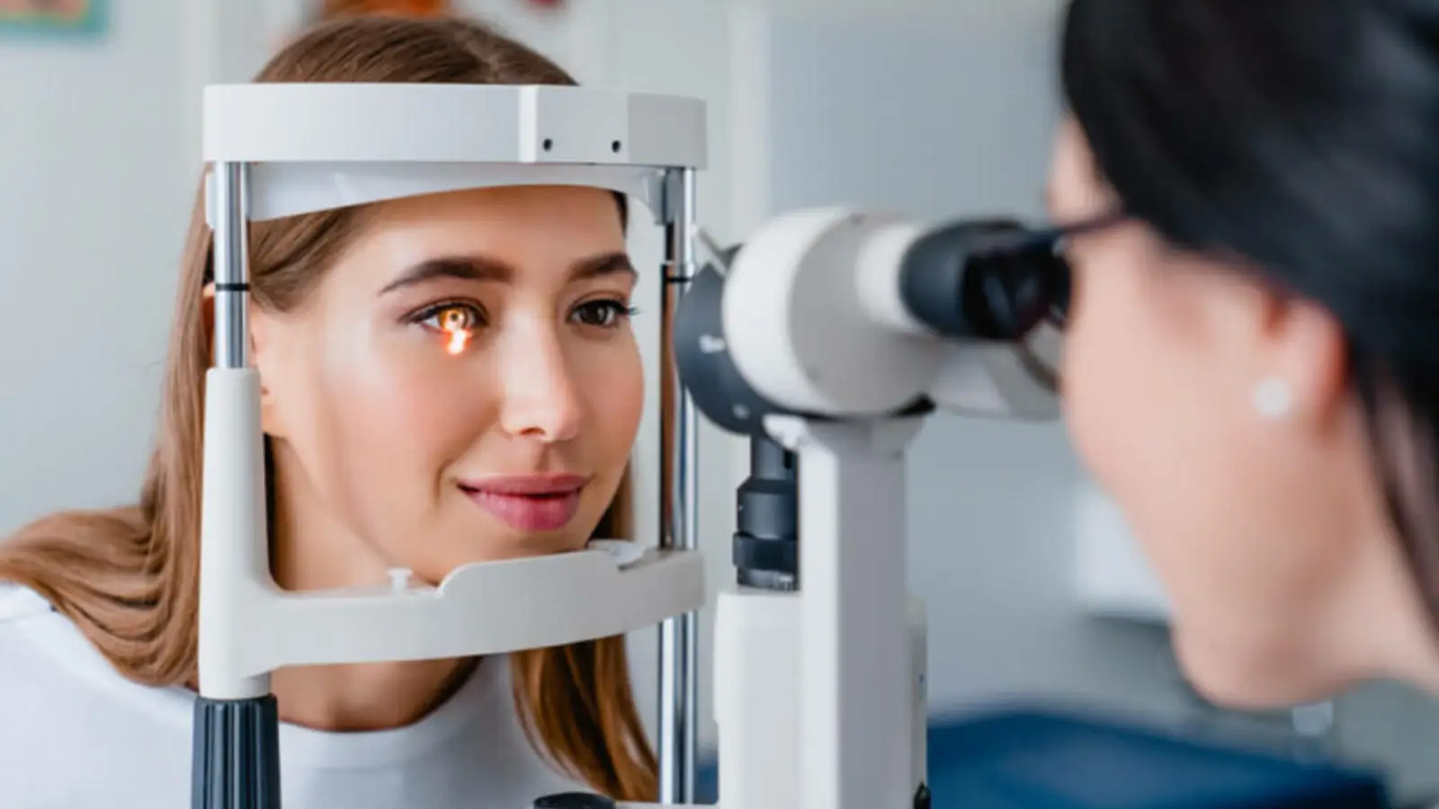 A woman is having an eye exam with a slit lamp. She rests her chin on the device, looking straight ahead as a healthcare professional observes her eye through the instrument. The background is a blurred clinical setting.