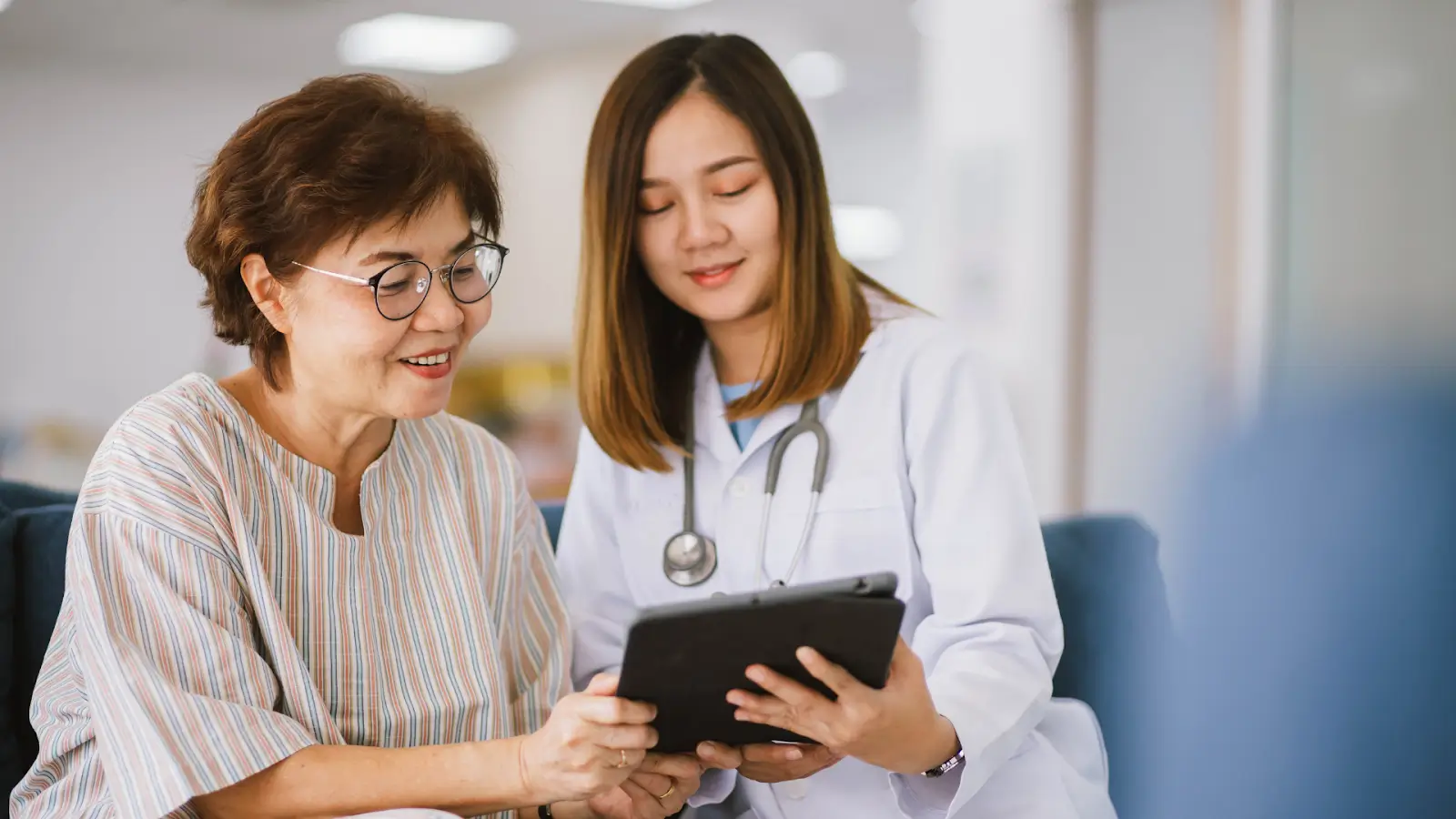 A doctor in a white coat, wearing a stethoscope, shows a tablet to a smiling older woman with glasses and short hair. They are seated indoors, engaged in a friendly conversation.