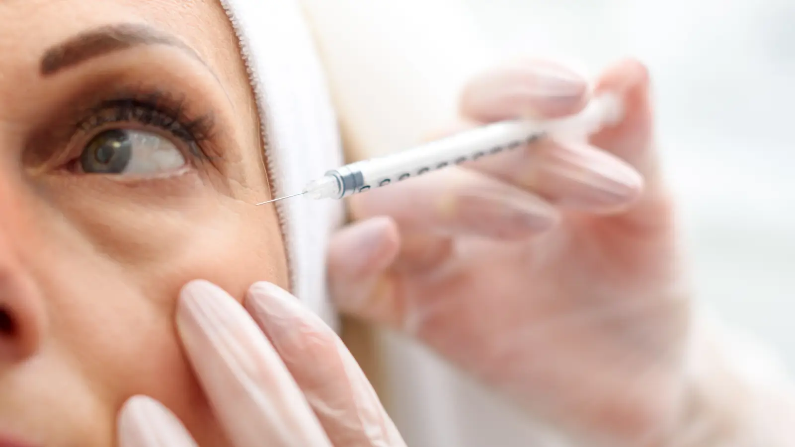 A close-up of a person receiving an injection near the eye. The person is wearing a white headband, and the hands holding the syringe are gloved.