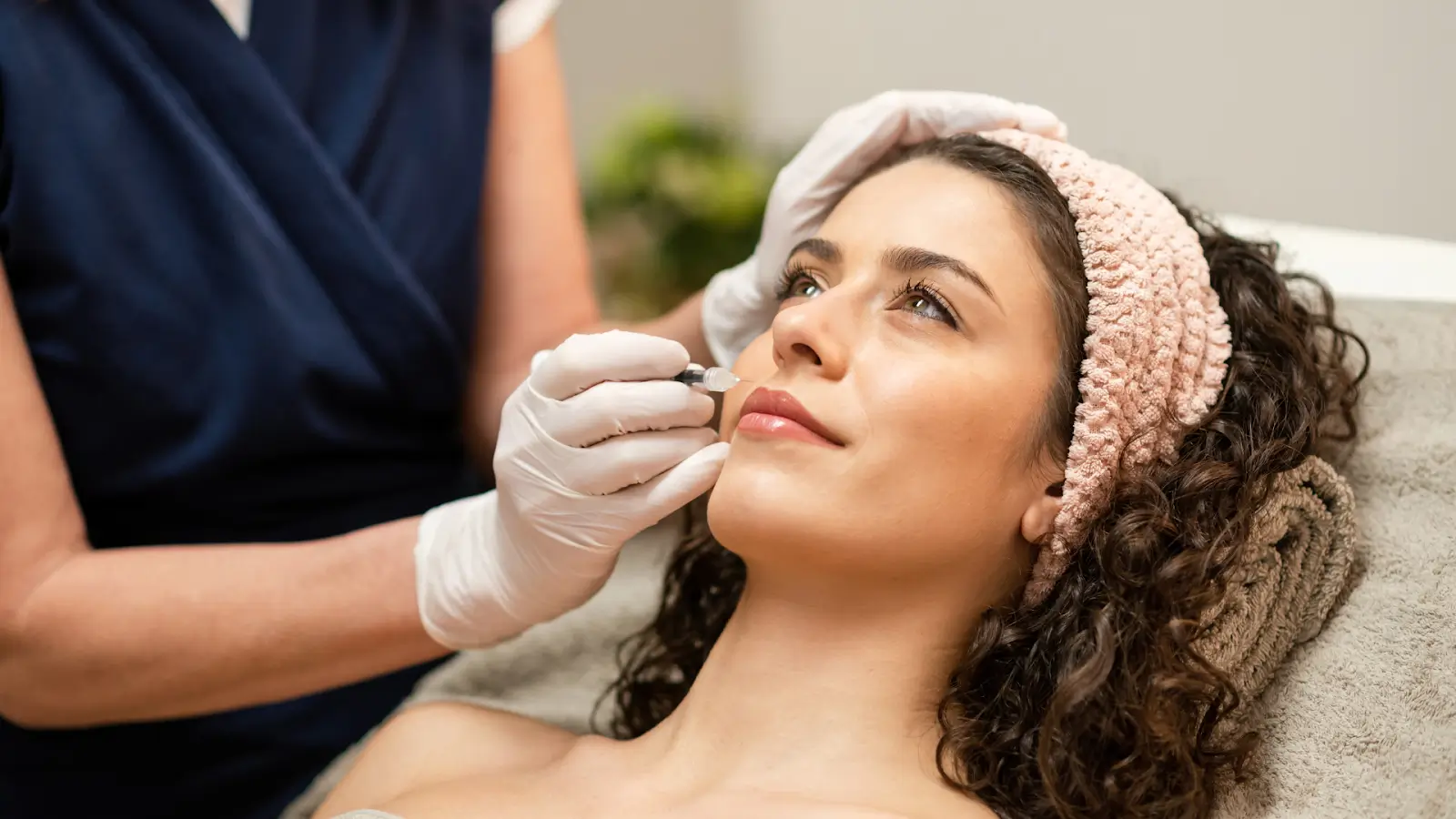 A woman with curly hair and a headband lies on a spa bed, receiving a lip treatment. A cosmetologist wearing gloves applies a product to her lips with a small tool.