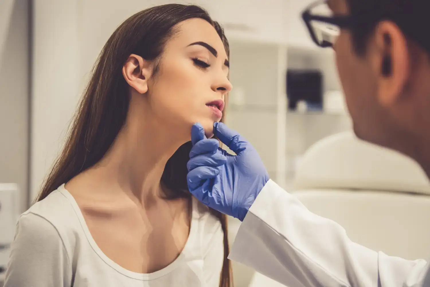 A doctor wearing blue gloves examines a woman's neck in a medical office. The woman, with long brown hair, is seated and looking upward slightly. Both appear focused during the examination. Medical equipment is visible in the background.