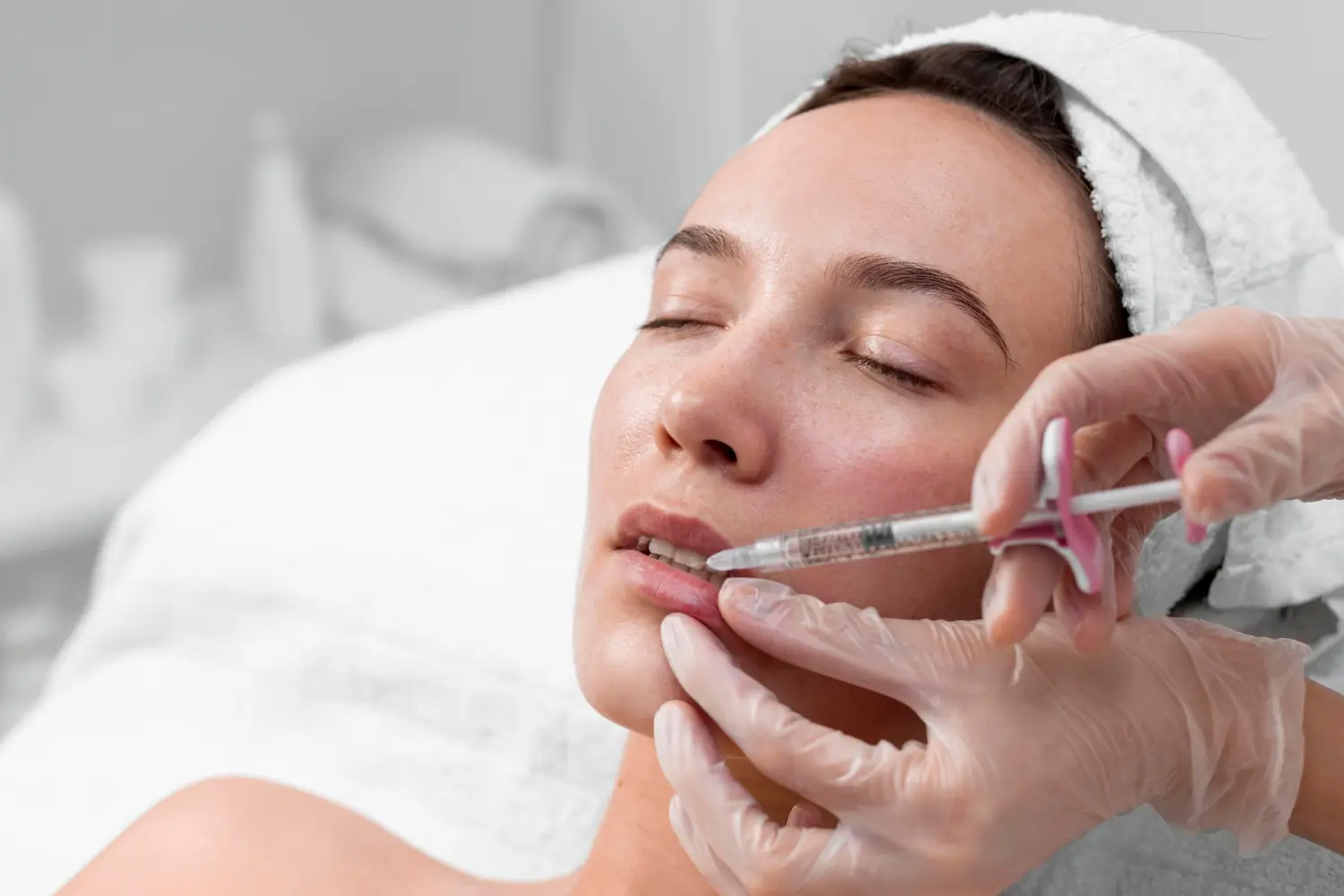 A person receiving a cosmetic injection in the lips. The individual is lying down with eyes closed, wearing a headband. The medical professional is using a syringe, wearing gloves, in a clinical setting.