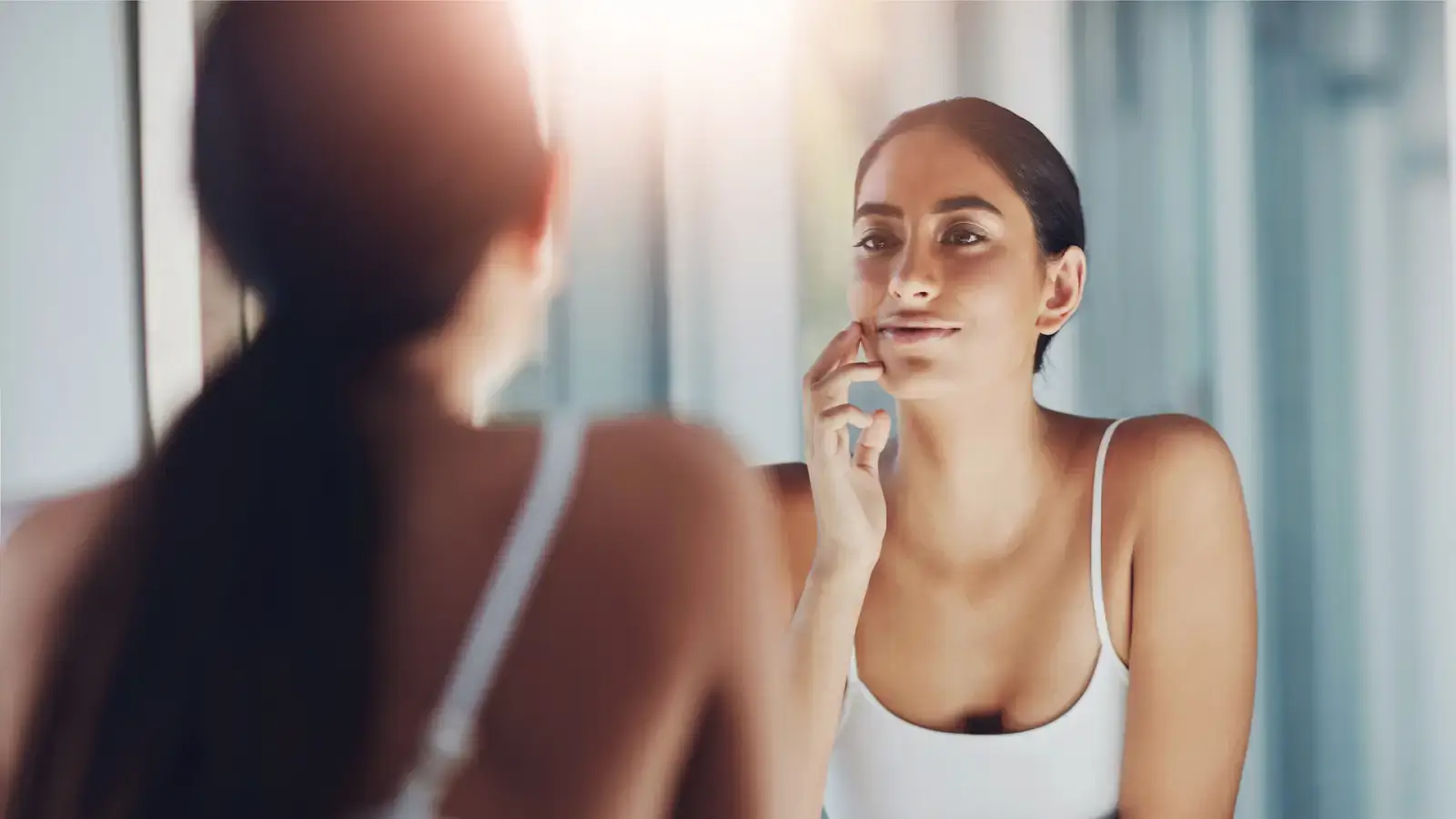 A woman in a white tank top looks at her reflection in the mirror with a soft smile, touching her cheek. The background is softly blurred with bright natural light.